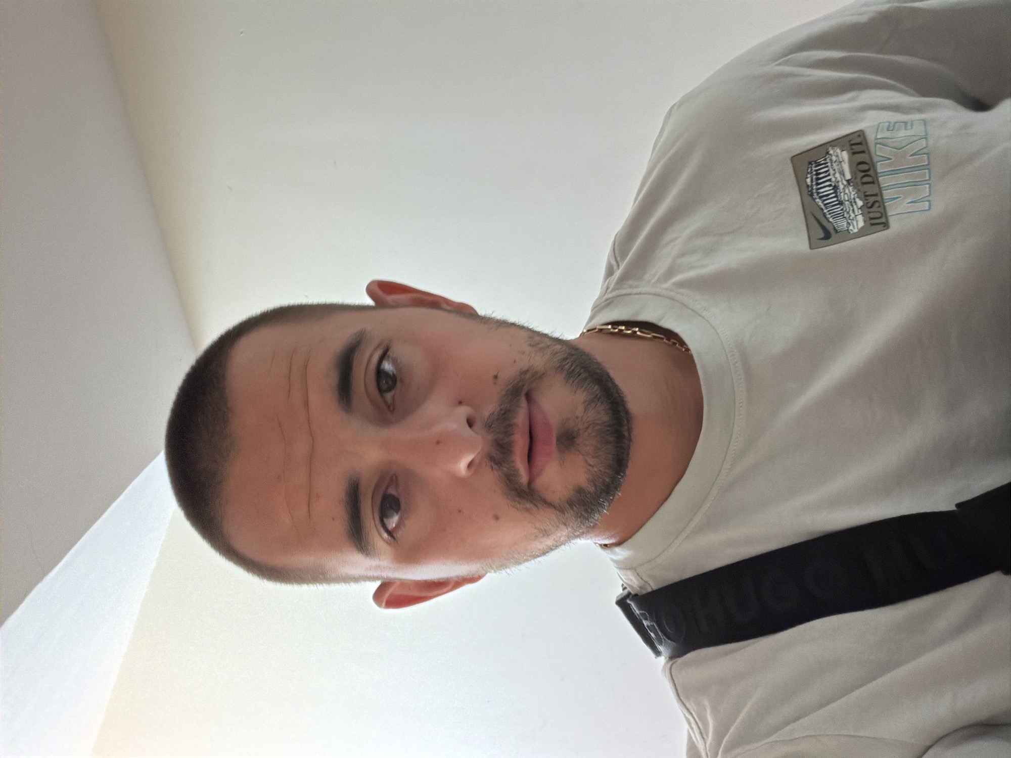 The image is a close-up, selfie-style portrait of a man with a buzz cut and a beard. He is wearing a light-colored t-shirt with a Nike logo and graphic on the left chest, and a black strap across his chest. He is also wearing a gold chain. The background appears to be an indoor setting with a white wall and ceiling. There is no identifiable information in the image that places it in Burgas, Bulgaria.
