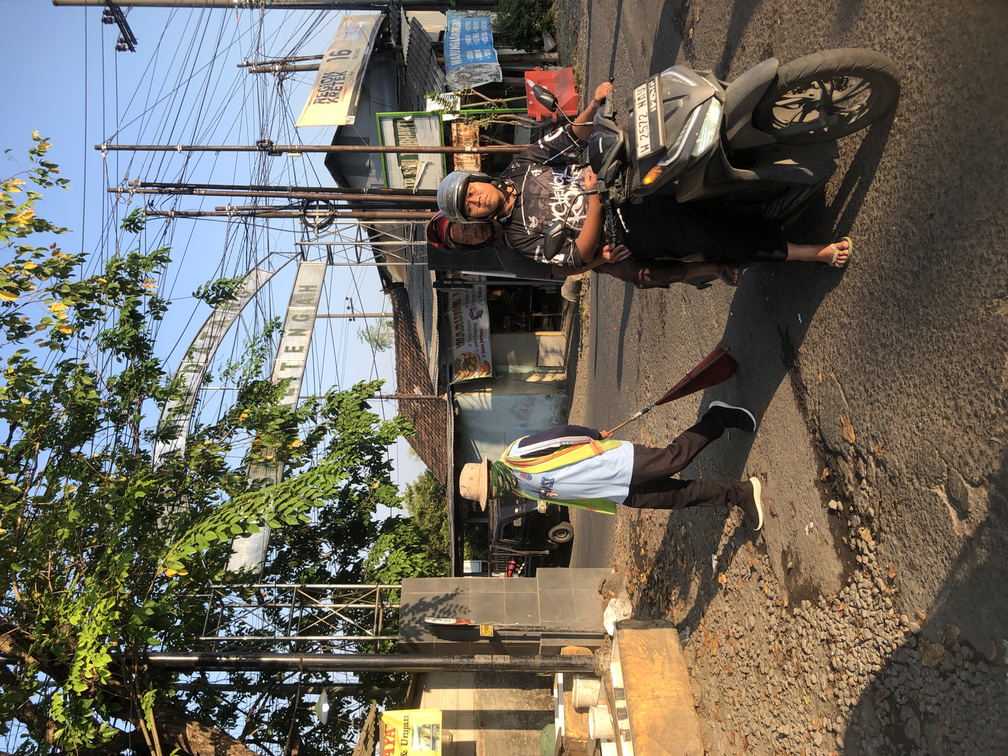 In Kludan, Indonesia, a street sweeper wearing a hat, a green face mask, a light blue long-sleeved shirt, a reflective yellow vest, and dark pants walks on the side of a road with a broom. He is heading towards the left side of the frame. Behind him, a motorcycle carrying two people rides down the road. The driver wears a helmet and a black shirt with a graphic design, while the passenger behind him wears a red helmet and a dark shirt. The motorcycle has a license plate that reads "W 2572 NAU". The environment appears to be a moderately developed area with buildings, power lines, trees, and a blue sky. A sign above an archway reads "DESA DUKUH". There are also several banners and signs visible on the buildings. The lighting suggests it is either early morning or late afternoon.