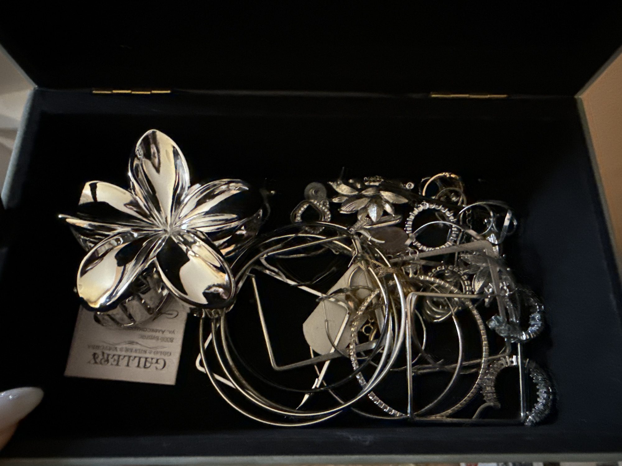 The image shows a dark jewelry box filled with silver-colored jewelry. The box appears to be lined with black fabric. On the left is a large silver flower-shaped brooch or hair clip with a white tag attached. Other pieces include multiple silver hoops, studs, and possible rings. The tag attached to the flower indicates “GALLERY”, but the rest of the address is illegible. The location of this is thus unknown.