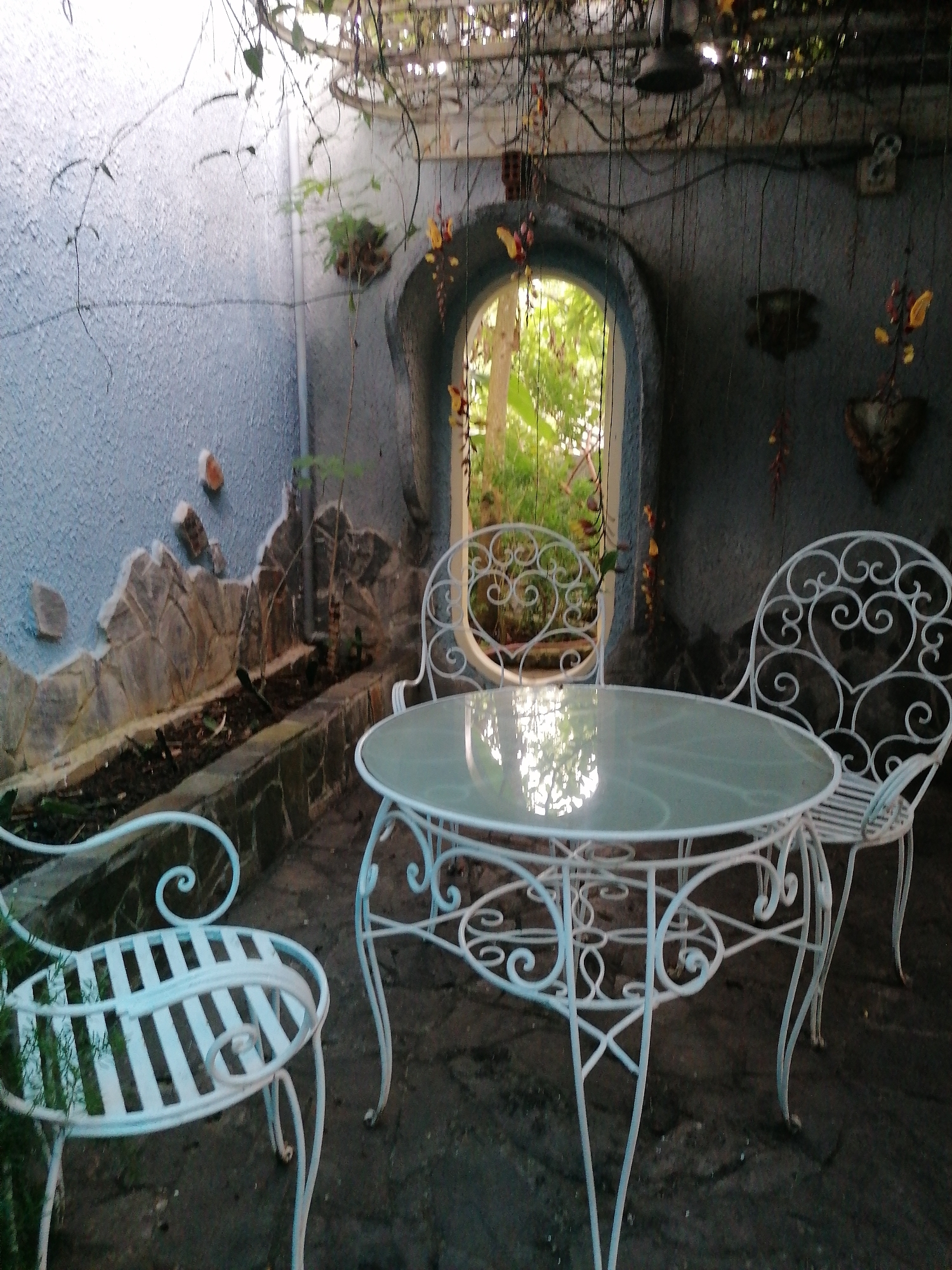This media file depicts an outdoor seating area in Da Lat, Vietnam. The scene features a round glass-top table with an ornate white metal base and matching white metal chairs, one of which is partially visible on the right. The furniture is set on a dark, possibly stone or concrete, patio.
Behind the table, an arched doorway leads to a glimpse of lush greenery and trees, suggesting a garden or natural setting. The walls surrounding the seating area are textured and painted a light blue. A decorative stone border runs along a planter box filled with soil and a few small plants. Overhead, vines and branches with yellow flowers hang down, creating a somewhat overgrown and bohemian atmosphere. A dark ceiling fan is visible attached to the structure above.
The lighting suggests it might be late afternoon or early evening, with soft natural light filtering through the doorway and dappled light from the overhead vines. The overall mood is peaceful and tranquil, with a hint of rustic charm. There is no visible text in the image.