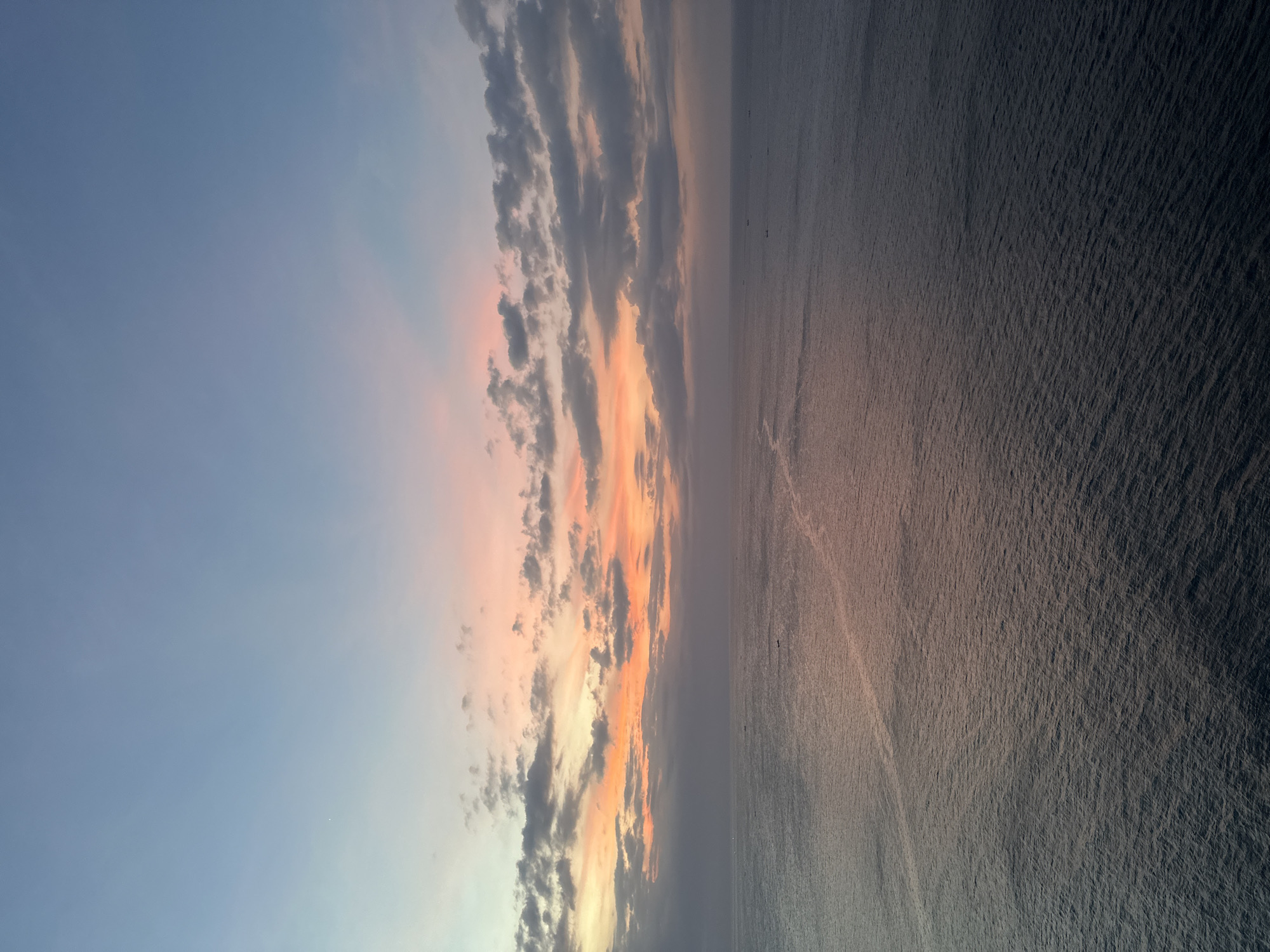 The image depicts a sunset view over the ocean in Kuta Selatan, Indonesia. The sky transitions from a pale blue to shades of pink and orange where the sun is setting. Clouds stretch across the horizon, illuminated by the setting sun. The ocean reflects the colors of the sky, with darker tones nearer the viewer and lighter, orange hues further out. A few small boats are visible on the water in the distance.
