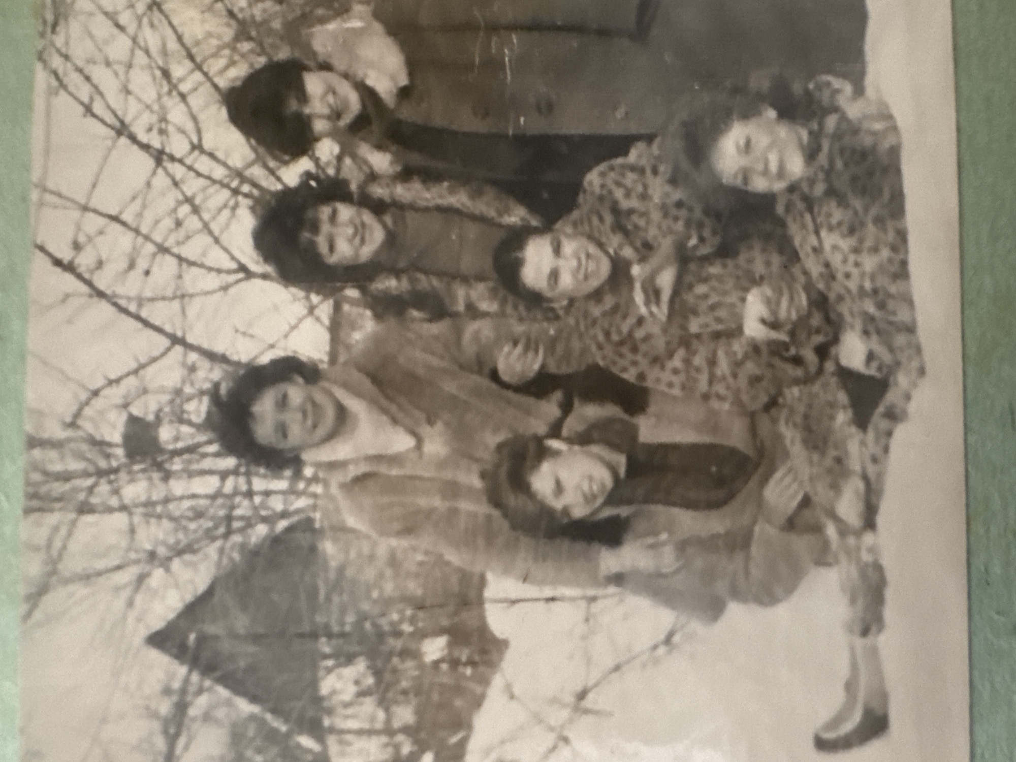 A black and white photograph shows six young women standing outdoors, possibly in a yard or park with bare trees in the background. The women are smiling and appear to be posing for the photo. One woman is wearing a patterned coat or dress, while others are wearing solid colored garments. One woman wears boots. No specific information in the image suggests a location in Ramat Gan, Israel.