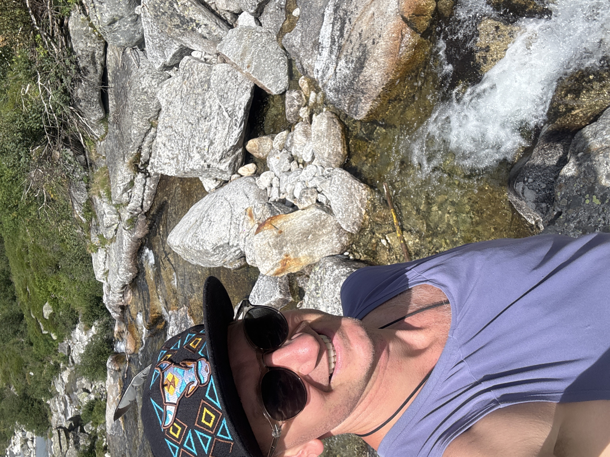 This is a portrait-style, high-angle, close-up selfie of a person standing near a rocky stream. The person is wearing a black baseball cap with colorful geometric patterns and an embroidered design of an animal's head, and round-lensed sunglasses. They are also wearing a light purple t-shirt and a black cord necklace. The person is smiling broadly at the camera. The background consists of large, grey rocks and a fast-flowing, clear stream with whitewater. Green vegetation is visible on the left side of the image, suggesting a natural, outdoor setting. The lighting indicates it is daytime, likely sunny given the bright conditions and shadows. The rocks and water suggest a mountainous or alpine environment. There is no visible text in the image. The overall mood is cheerful and adventurous.