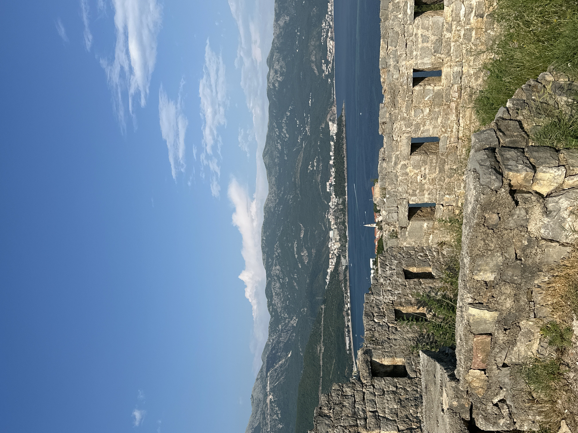 The image captures a landscape view from the medieval fortification of Stari Grad, or Old Town, in Budva, Montenegro. A weathered, light-colored stone wall with rectangular openings occupies the foreground. Beyond the wall, a cityscape stretches along the coast, nestled between the Adriatic Sea and a backdrop of mountains. The sky is a bright blue with scattered white clouds.