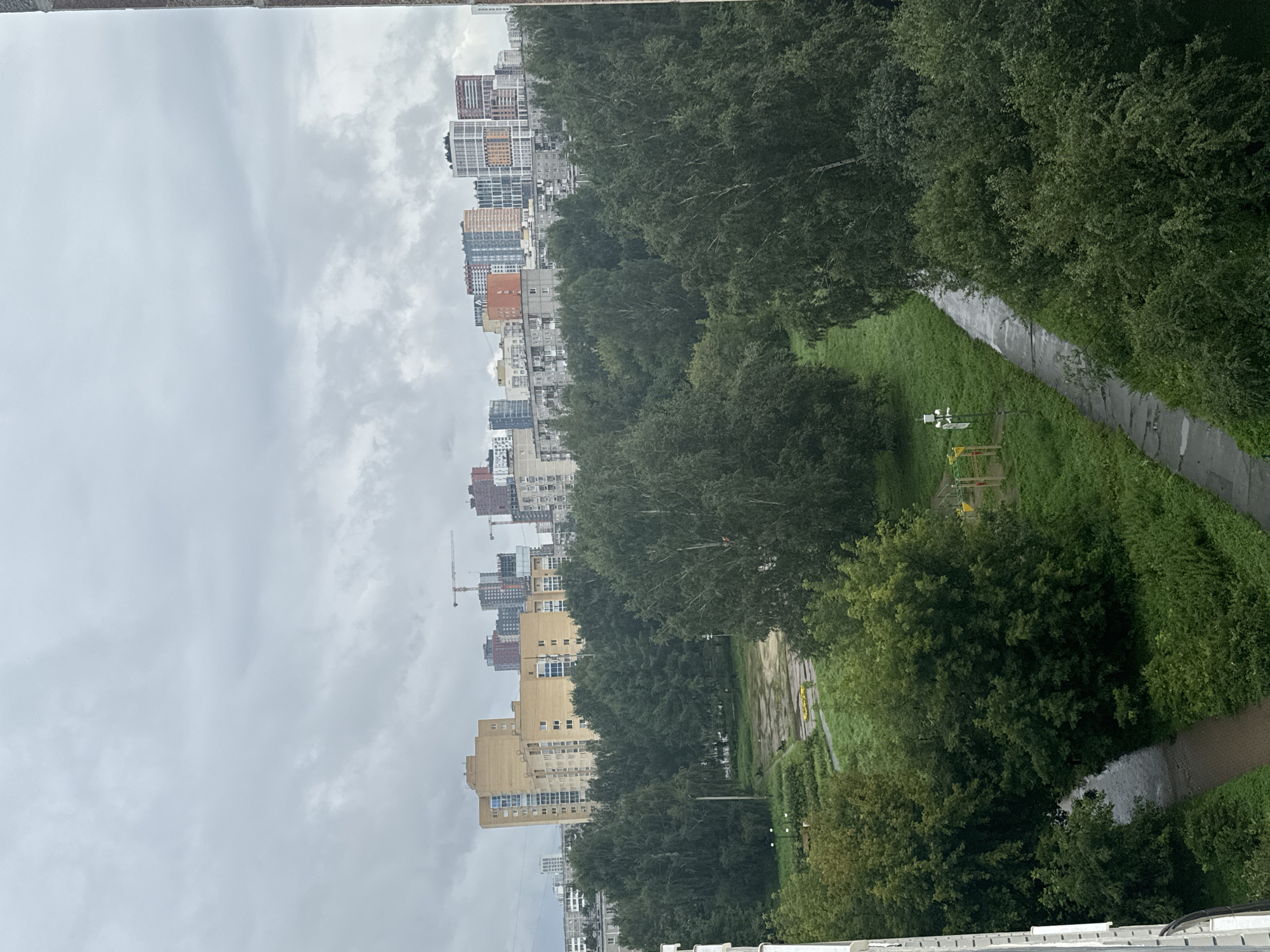 The image captures an overcast day in a city, likely in the suburbs or a residential area. In the background, a cluster of tall buildings, ranging in color from beige to vibrant hues of orange, pink, and blue, punctuate the skyline. Construction cranes are visible among some of these structures, suggesting ongoing development. Closer to the foreground, a dense canopy of mature green trees dominates the landscape. Within the treeline, a paved path winds through lush grass, bordered by more trees. On the left side of the path, a small playground with yellow and red structures is visible, along with a lamppost. The ground appears damp in places, possibly indicating recent rain. The overall scene depicts a blend of urban development and natural greenery under a grey, cloudy sky.