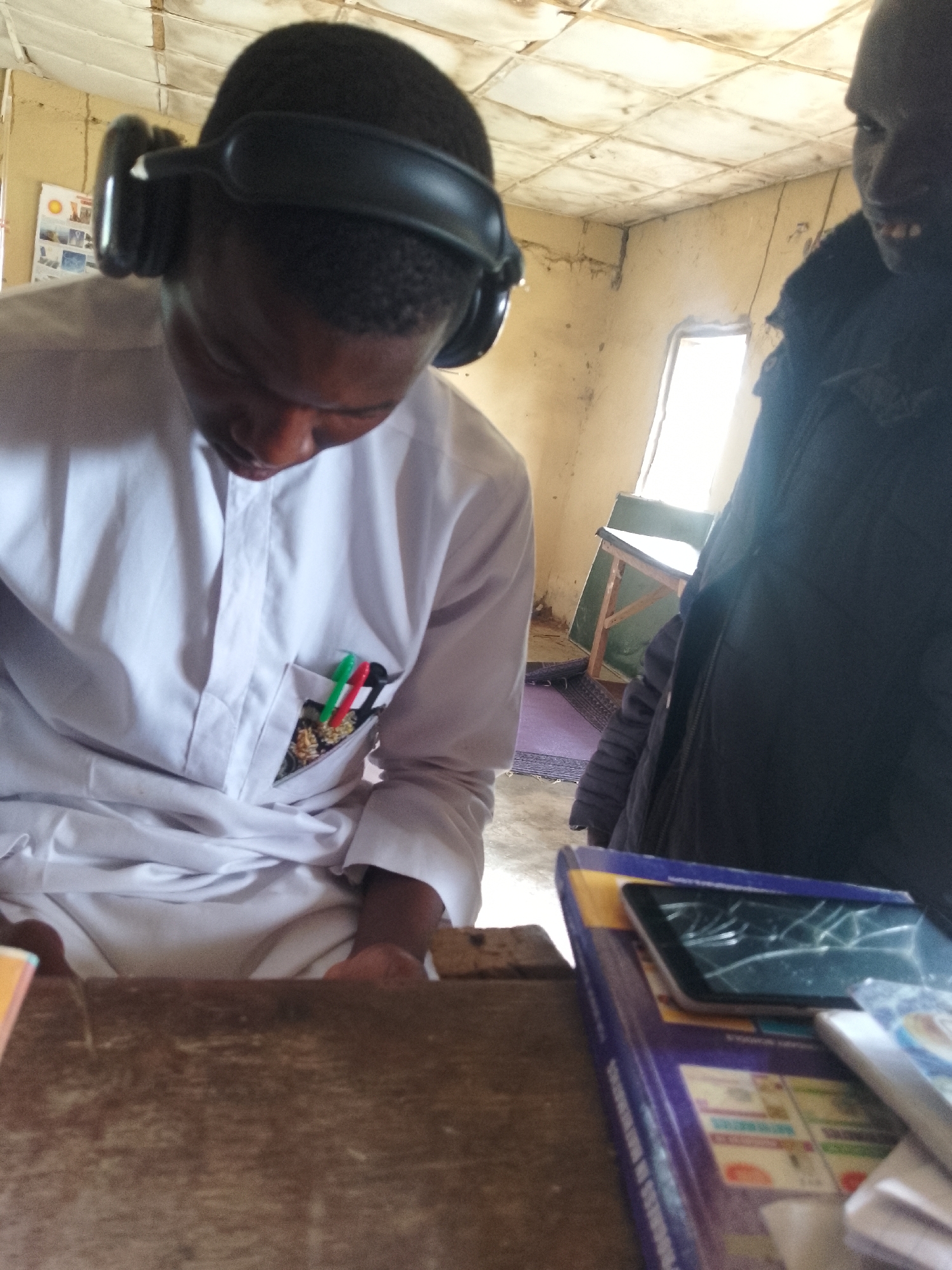 In a classroom in Serti, Nigeria, a young boy wearing headphones is seen looking down at a desk. He is wearing a white tunic with pens in the pocket. To the right, another person, partially obscured, appears to be looking at the boy. The room has a tiled ceiling and a window is visible in the background. On the desk in front of the boy is a tablet with a cracked screen, placed on top of some books. A wooden desk is in the foreground.