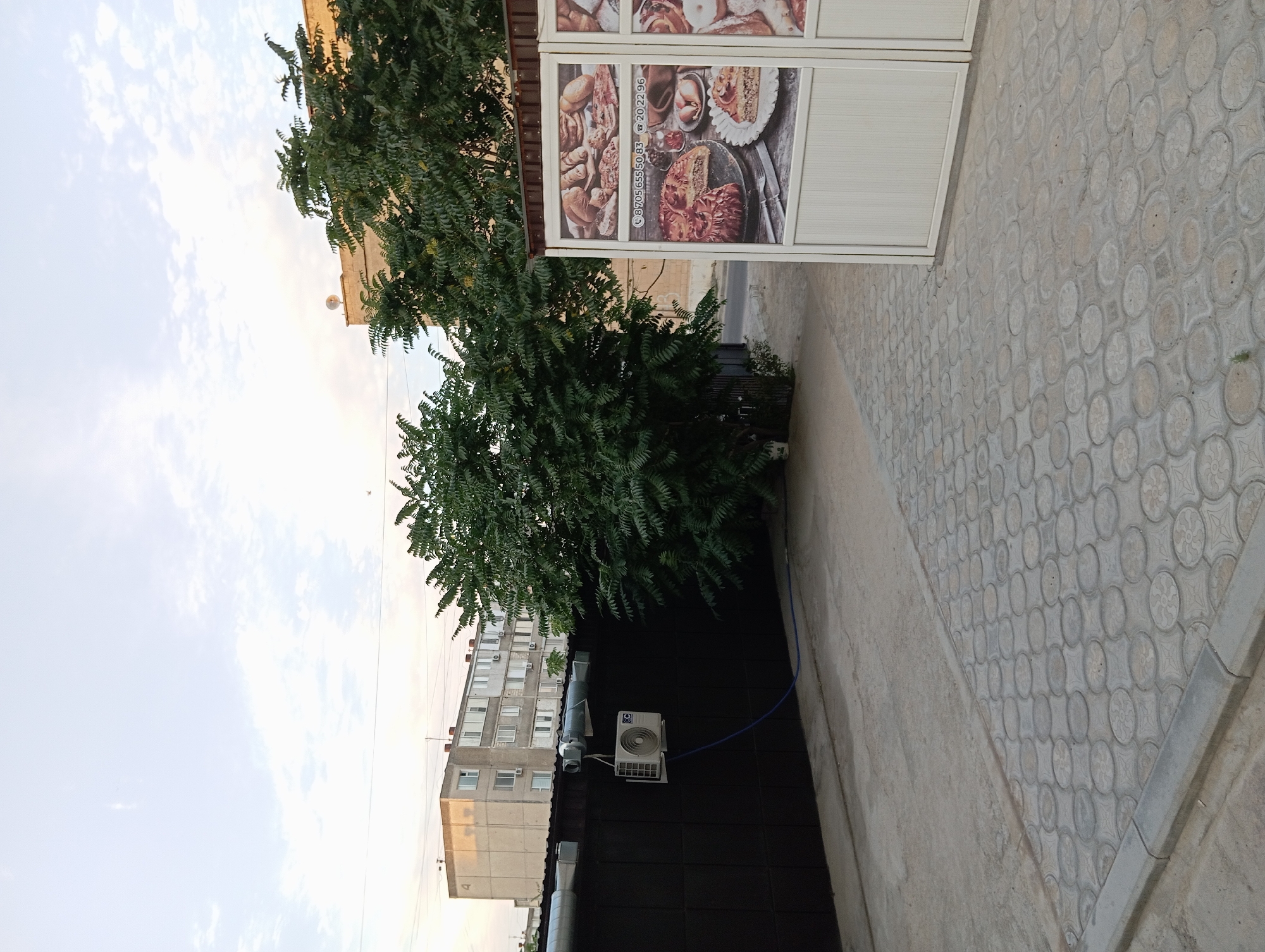 The image shows an outdoor scene in Aktau, Kazakhstan. In the background, a multi-story apartment building is visible, with a concrete fence in front of it. An air conditioning unit is mounted on the fence, with a blue hose connected to it. To the right, a structure with advertising panels featuring images of baked goods is present. A large tree with dense green foliage stands between the fence and the advertising structure. The ground is paved with concrete and decorative tiles. The sky is partially cloudy.