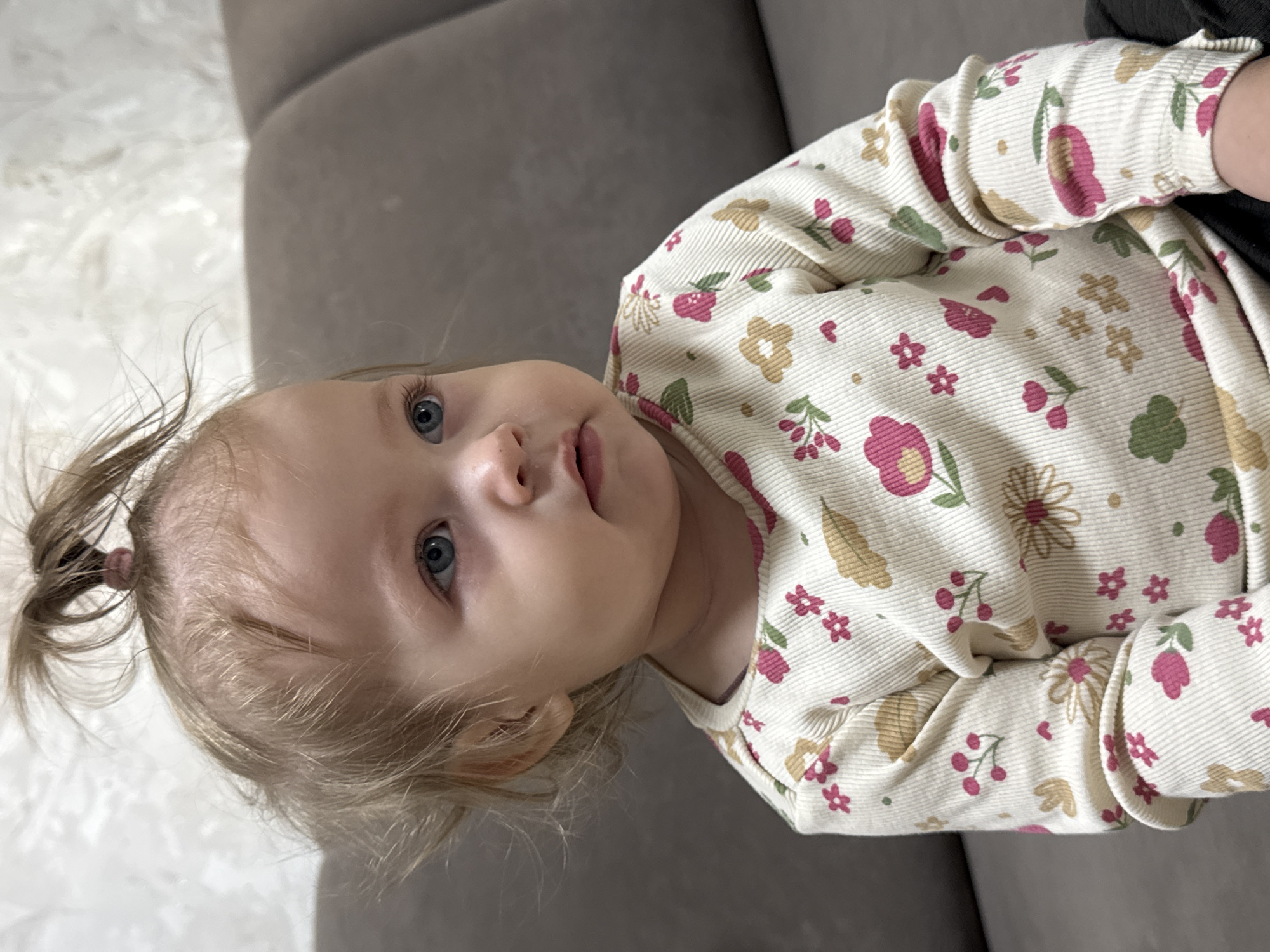 A young child with light blonde hair and blue eyes is lying on a grey sofa. The child wears a long-sleeved, patterned shirt with floral motifs in pink, green, and tan. The shirt's fabric appears to be corduroy. The child's hair is partially tied up in a small ponytail. The background includes a section of the grey sofa and, out of focus, a light-colored surface possibly a carpet or another section of the sofa. There's no information in the image about the location being in Chaikovsky, Russia.