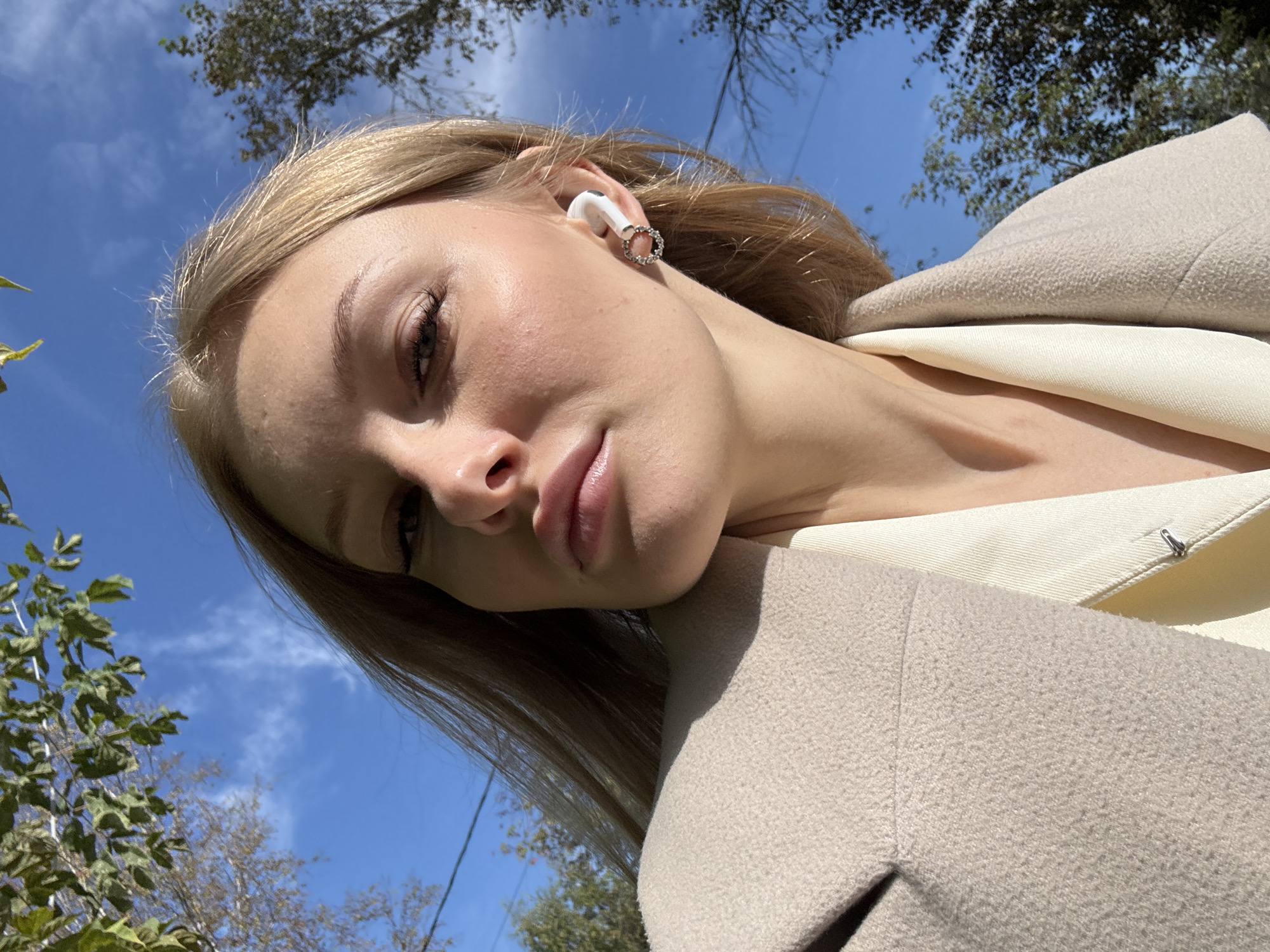 A young woman with blonde hair and fair skin is shown in a close-up selfie taken outdoors. She is wearing a light-colored coat and a cream-colored shirt. An AirPod is visible in her right ear, and she is wearing a decorative earring on the same ear. The background consists of a bright blue sky with some clouds, and the tops of trees with green and brown leaves are visible. The lighting suggests it is daytime. The image is a selfie, likely taken by the subject herself, pointing the camera upwards. No specific landmarks within the city of Perm, Russia, are identifiable in this image.