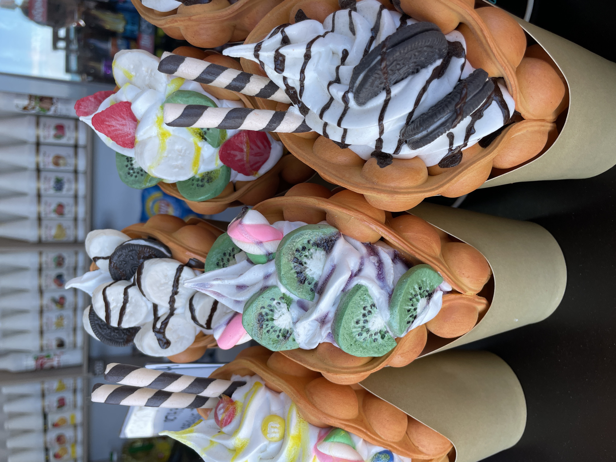 The image displays a close-up, overhead view of four vibrantly decorated dessert cones, likely a variation of Hong Kong-style egg waffles or bubble waffles, presented in individual beige paper holders. These cones are arranged side-by-side in what appears to be a food stall or dessert shop display, with the image rotated 90 degrees clockwise from a typical upright view.
Each golden-brown, bubbly waffle cone is generously filled with white whipped cream or soft-serve ice cream and a variety of sweet toppings.
The uppermost cone features whipped cream drizzled with chocolate syrup, topped with two Oreo cookies and two striped chocolate and vanilla wafer rolls.
Below this, the next cone is adorned with sliced green kiwi fruit and small pink and white marshmallow candies, with a hint of purple swirl in the cream.
The third cone showcases a colorful assortment of sliced red strawberries, sliced yellow bananas, and more green kiwi slices, along with a yellow drizzle and two striped wafer rolls.
The bottom-most cone is topped with white cream, various colorful candy pieces (yellow, green, pink, blue), and a striped wafer roll.
The background is significantly blurred, showing reflections in a glass surface and indistinct shelving units with unidentifiable products, suggesting an indoor or semi-outdoor commercial setting. The lighting indicates it is daytime. No specific activity is captured, but the presentation implies the desserts are ready for sale or consumption. There is no legible text visible within the frame. The desserts are fresh and appealing, highlighting their varied textures and toppings.