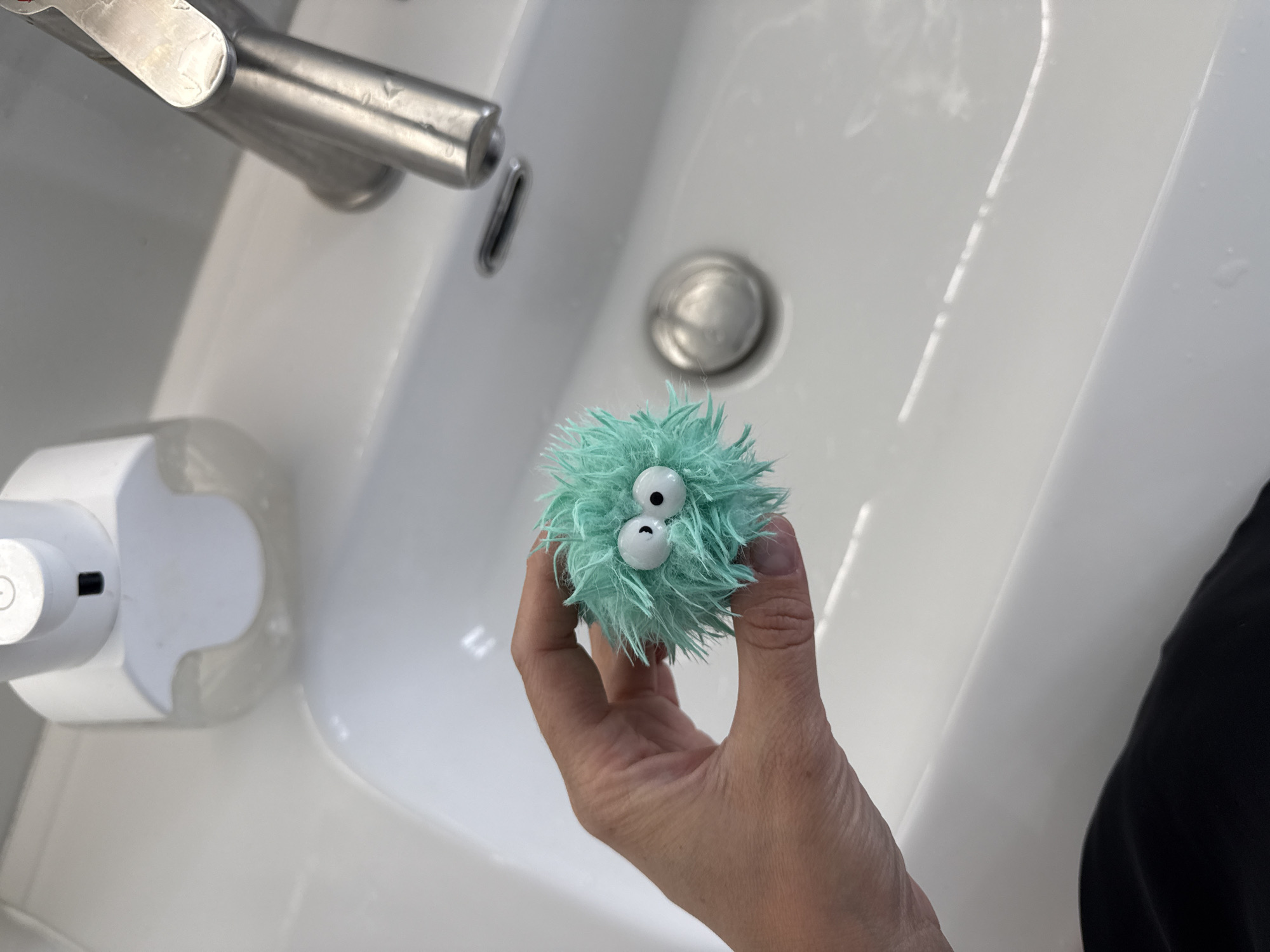 A close-up, high-angle shot shows a person's hand holding a small, fuzzy, teal-colored creature with large googly eyes over a white ceramic sink. The creature resembles a dust bunny with an expressive face. To the left of the hand is a white automatic soap dispenser. To the right, a silver faucet is visible, and further to the right, the edge of the sink curves away. The sink itself appears clean and wet, with reflections on its surface. The background is out of focus, but hints at a bathroom setting. The image was taken in Nizhneyarkeyevo, Russia.