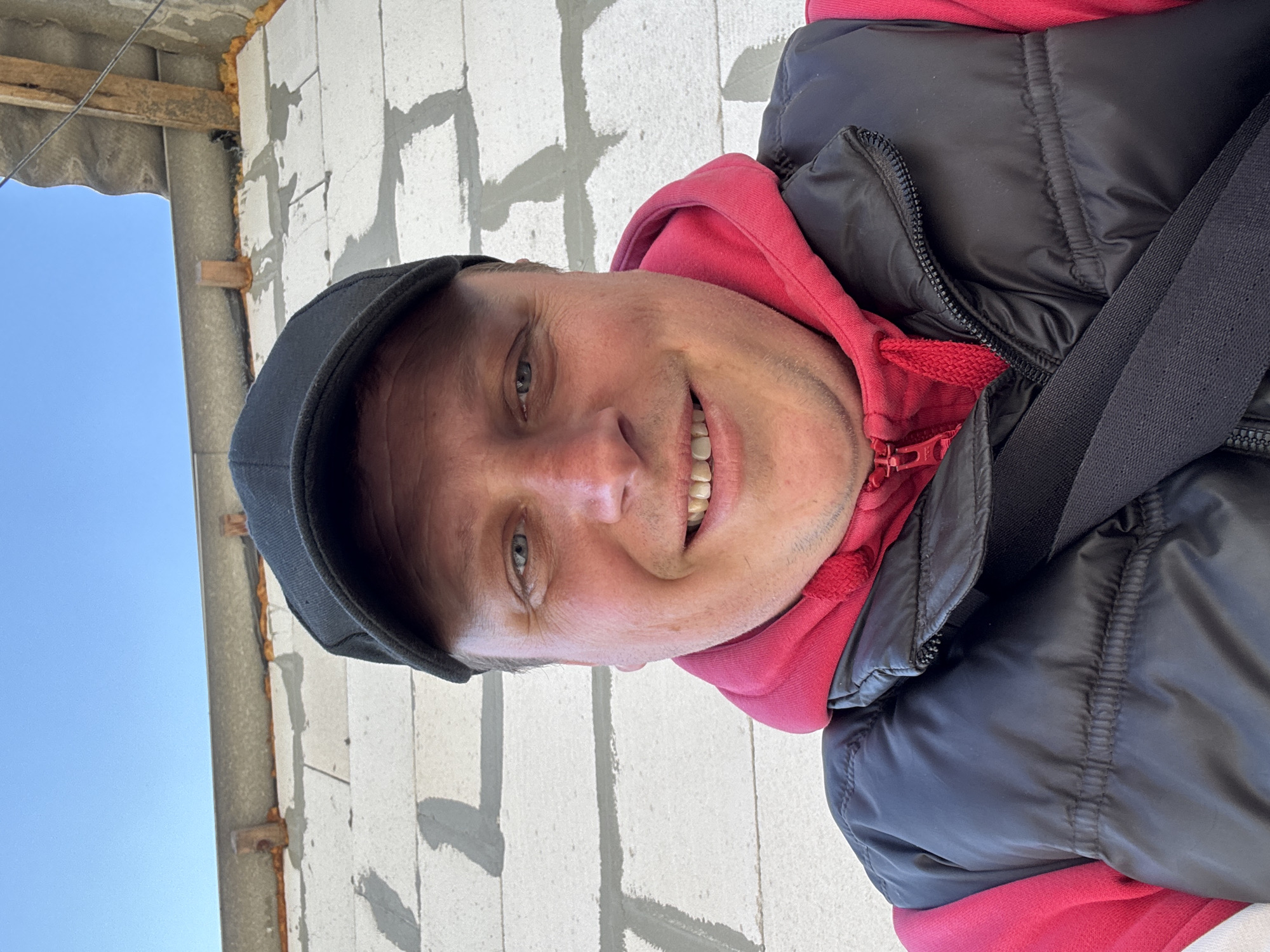 A man wearing a black cap, red hoodie, and black vest is smiling and looking directly at the camera. He is standing in front of a white brick wall under construction, with some of the bricks mortared together. The top left of the frame shows an open area with a blue sky visible. The location is Unknown, Unknown.