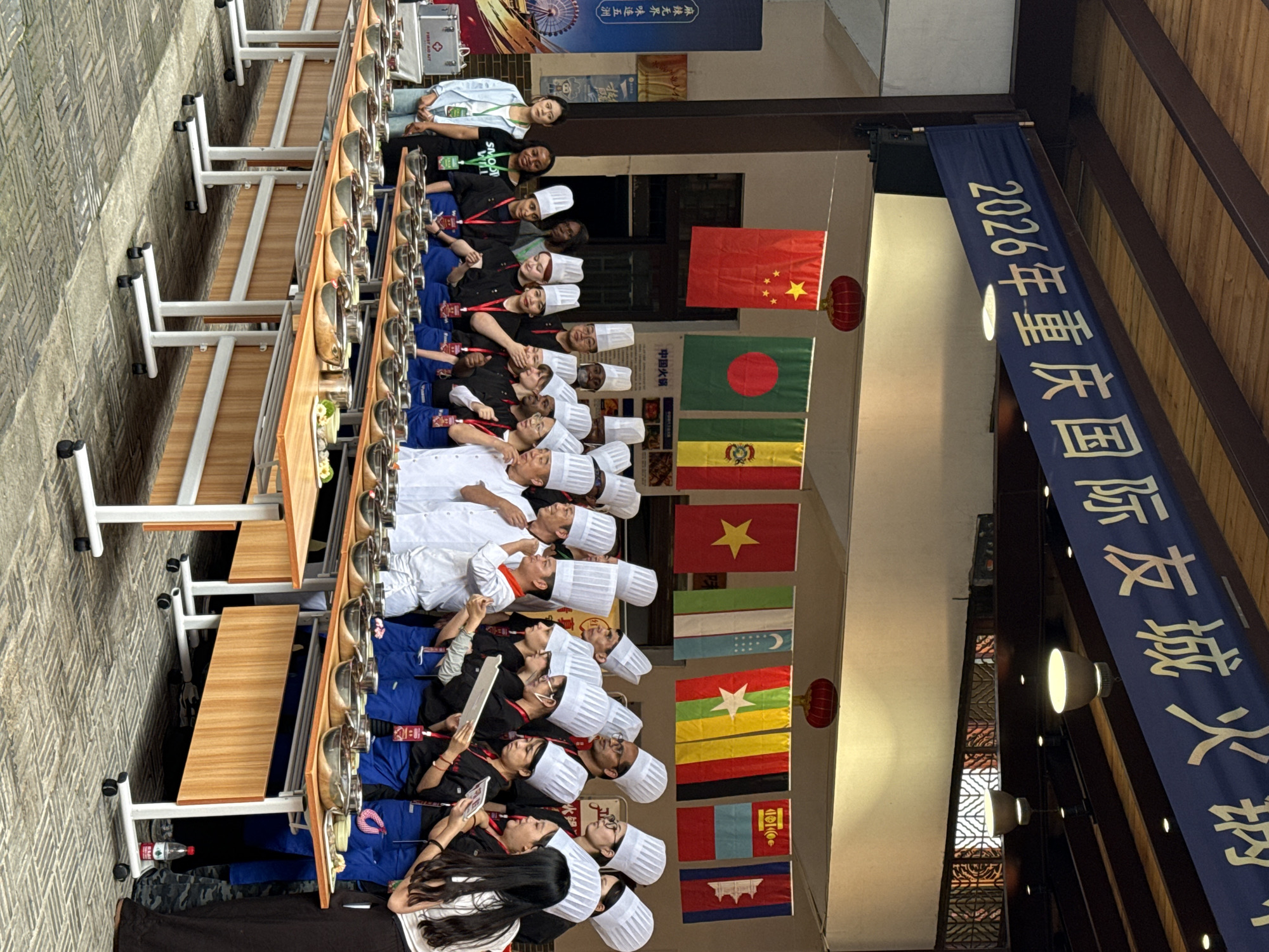 A horizontal shot depicts a group of people, primarily chefs in white hats, seated at tables with individual hot pot setups, under a display of international flags and banners, likely in Nan'an District, China.
The people are arranged in rows, facing the same direction, and appear to be participating in some sort of event or competition. The chefs are wearing uniforms with white chef hats and some are using tablets or paper.
Above them, hanging from the ceiling, are a series of national flags, including those of China, Bangladesh, and Vietnam. A blue banner with Chinese characters reading "2026年重庆国际友城火钢" (likely related to an event or festival in Chongqing) is also visible. The text "麻辣无界 味连五洲" (implying the spicy flavors of hot pot connect the world) and "SMOOT WILL" is displayed on a nearby wall.
The tables are lined with individual hot pot setups, indicating that the event involves cooking or food preparation.