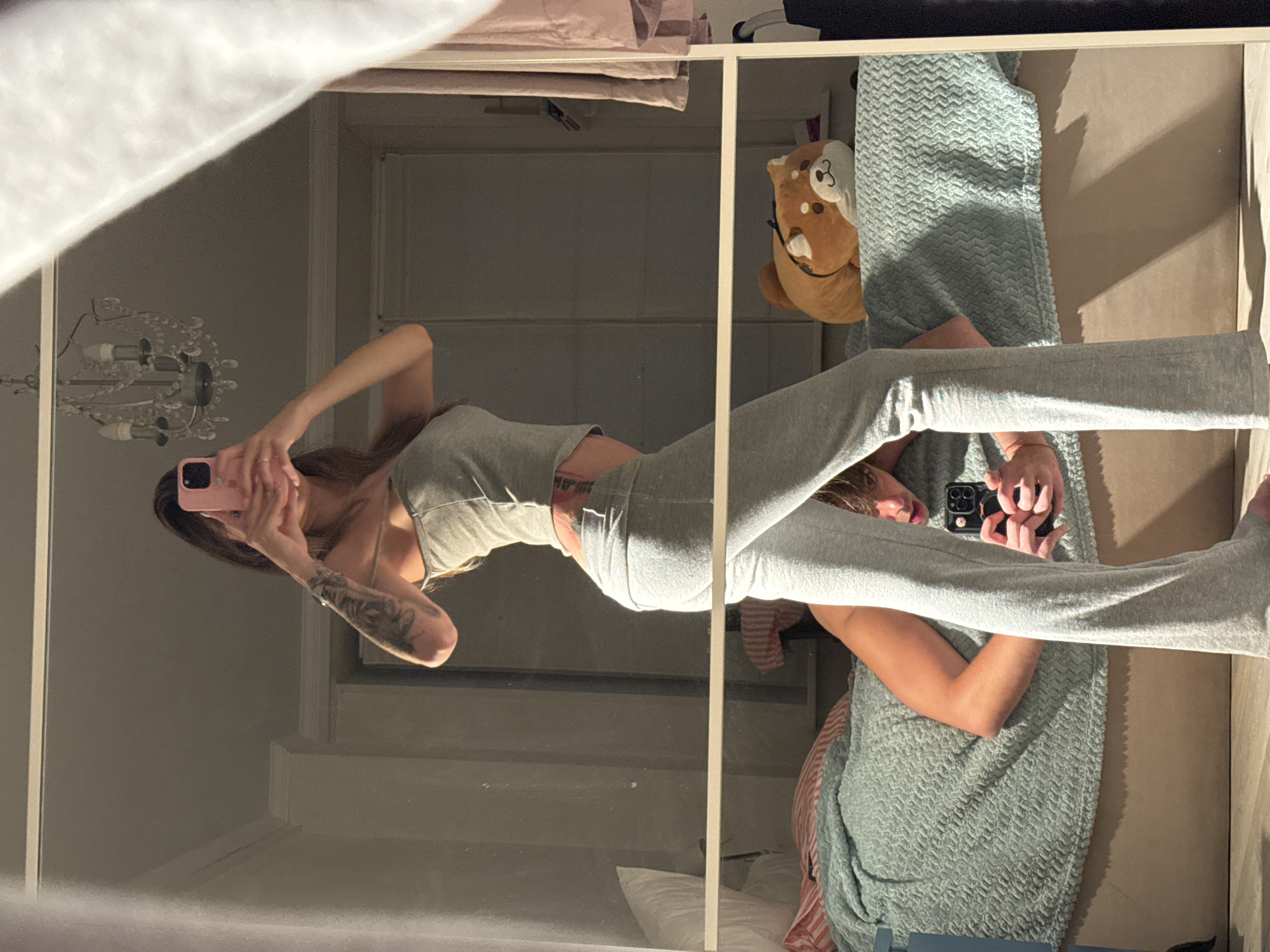 In a bedroom setting, a woman with long brown hair is taking a selfie in a mirror. She is wearing a grey tank top and grey sweatpants. She has tattoos on her left arm and lower back. A man is visible lying on the bed behind her, holding a phone and looking at the camera. A shiba inu plush toy is on the bed. The room features a chandelier and curtains. The image does not contain any information to suggest it is located in Moscow, Russia.
