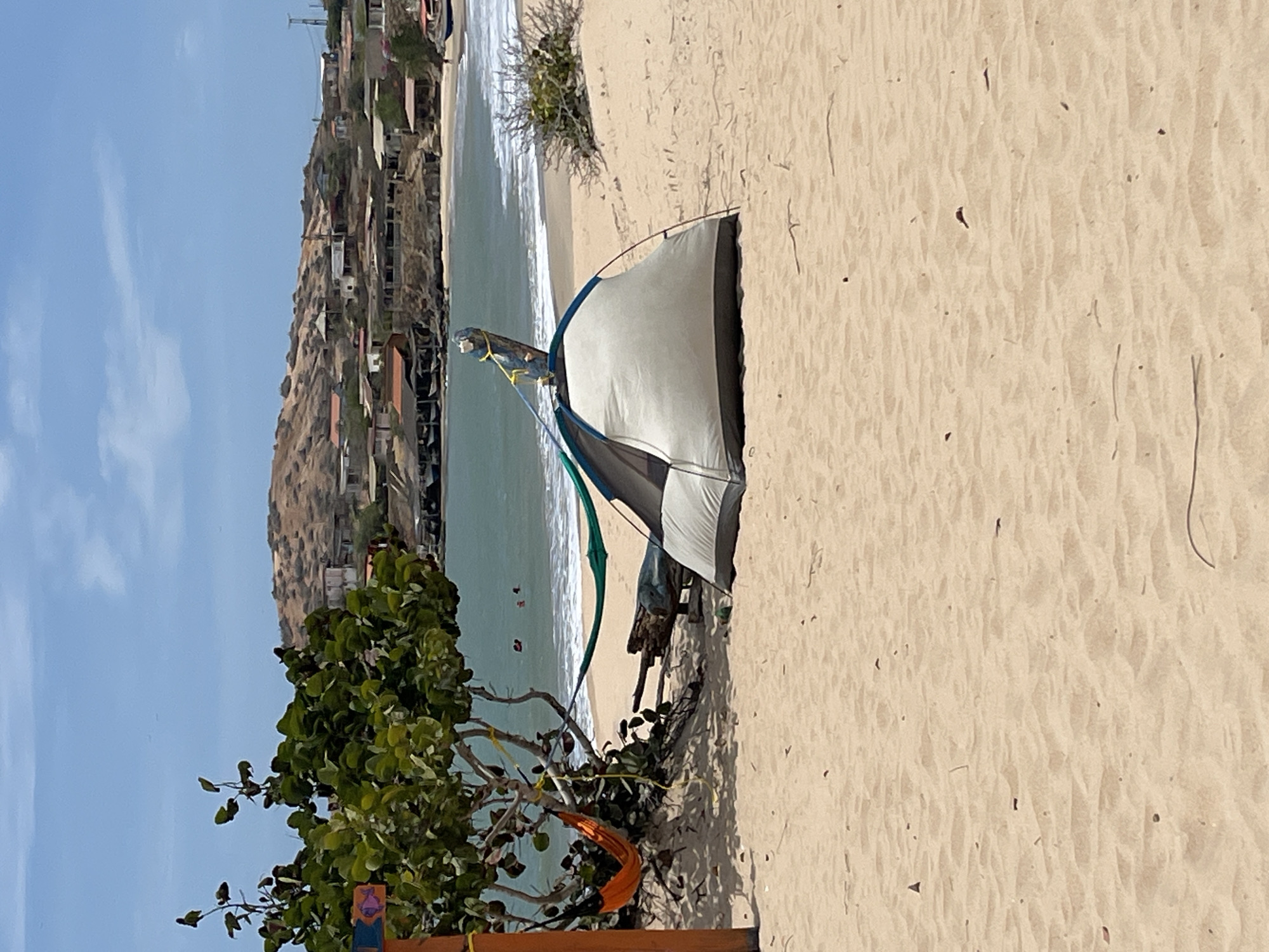 The image captures a beach scene in what appears to be La Pared, Venezuela, based on the presence of buildings on the hillside in the background. A light-colored tent is erected on the sandy beach, adjacent to some vegetation. There are people visible in the water, likely swimming or bathing. An object with orange fabric is attached to some branches. The environment is sunny.