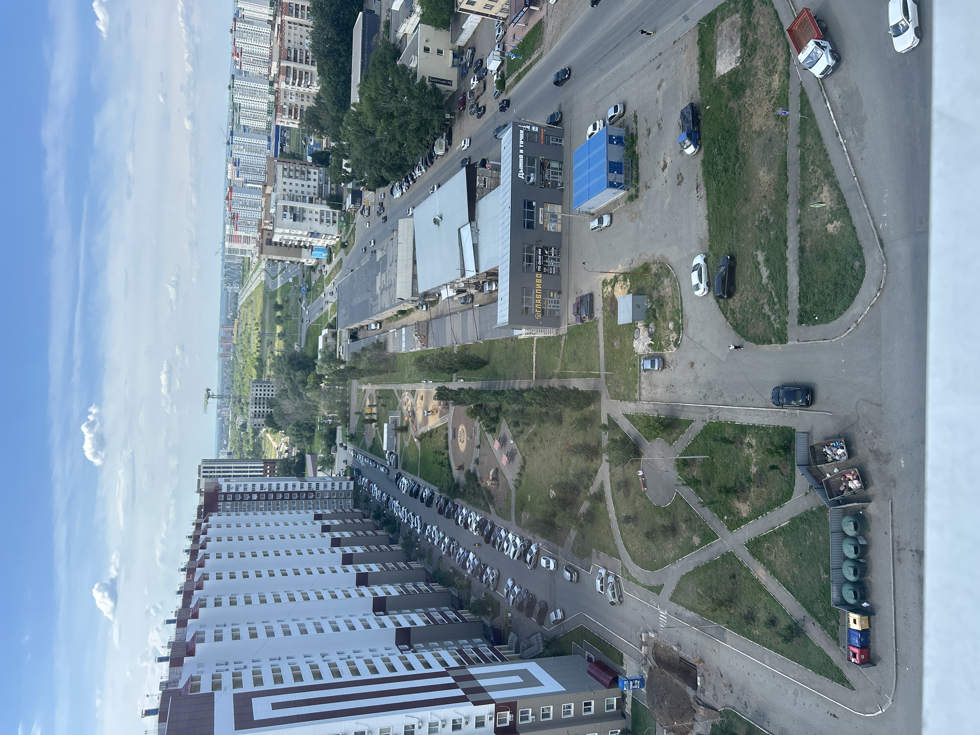 This is an aerial view of a residential area in Orenburg, Russia, likely taken from a tall building. The scene depicts a street lined with apartment buildings and commercial structures, with a clear blue sky and scattered white clouds indicating fair weather during the day.
The foreground is dominated by a long, multi-story apartment building with a white facade and dark red accents. Adjacent to it, a smaller commercial building features a dark grey exterior with signage that includes Cyrillic text, such as "ДЫМОМ И ТОЧКА" and "ПРАВИЗВО". A blue-roofed structure, possibly a canopy or small building, is situated near the commercial area.
The street itself is filled with parked cars of various colors and models, predominantly white, black, and grey. A few cars are in motion. A playground with colorful equipment is visible within a green, grassy area between the apartment buildings and the street. Further down the street, a row of dumpsters, some open and filled with refuse, and several green cylindrical bins are lined up. A white truck with a red flatbed is parked nearby, and other vehicles, including a black SUV and a white passenger car, are also present.
In the background, more apartment buildings and trees stretch into the distance, with a construction crane visible near some of the taller structures. The overall impression is of a typical urban residential and commercial environment on a sunny day. No people are clearly discernible, suggesting the photo was taken from a distance or at a time with minimal pedestrian activity.