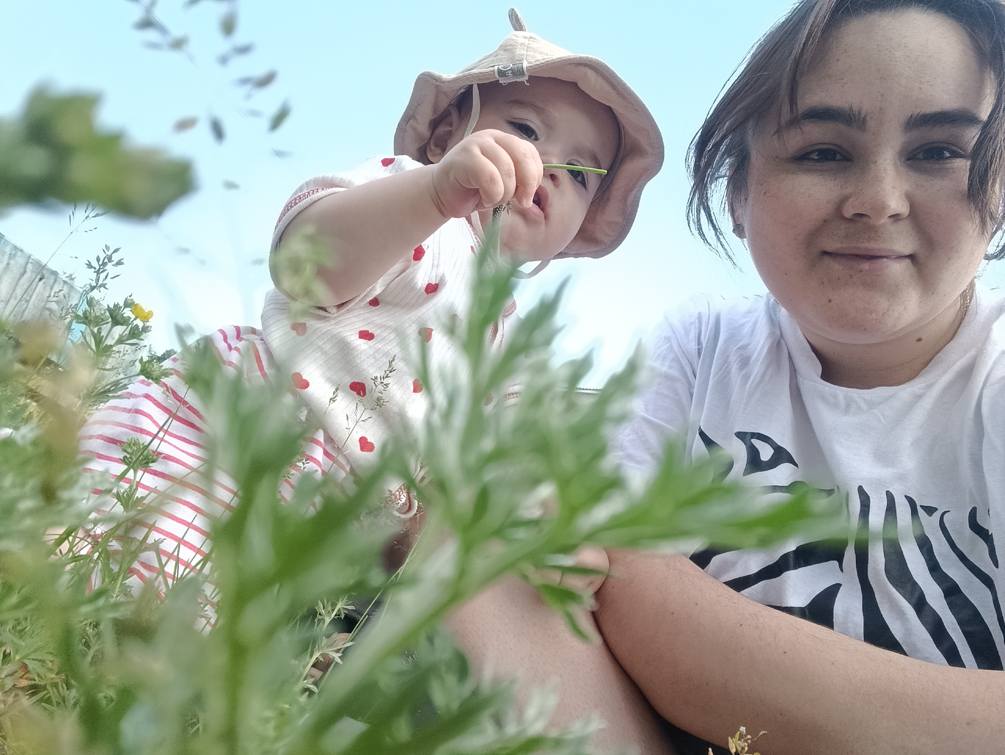 A low-angle, close-up selfie shows a woman and a baby outdoors in the city of Noviy, Russia. The woman, in the foreground on the right, wears a white t-shirt with a black zebra stripe pattern. She is looking directly at the camera with a slight smile. To her left and slightly behind her, a baby wearing a light-colored bucket hat and a white onesie with red hearts and red and white striped pants is reaching out and touching a blade of grass. The background is a bright blue sky with some blurred foliage and a wooden fence on the left. The overall scene suggests a casual outdoor activity.