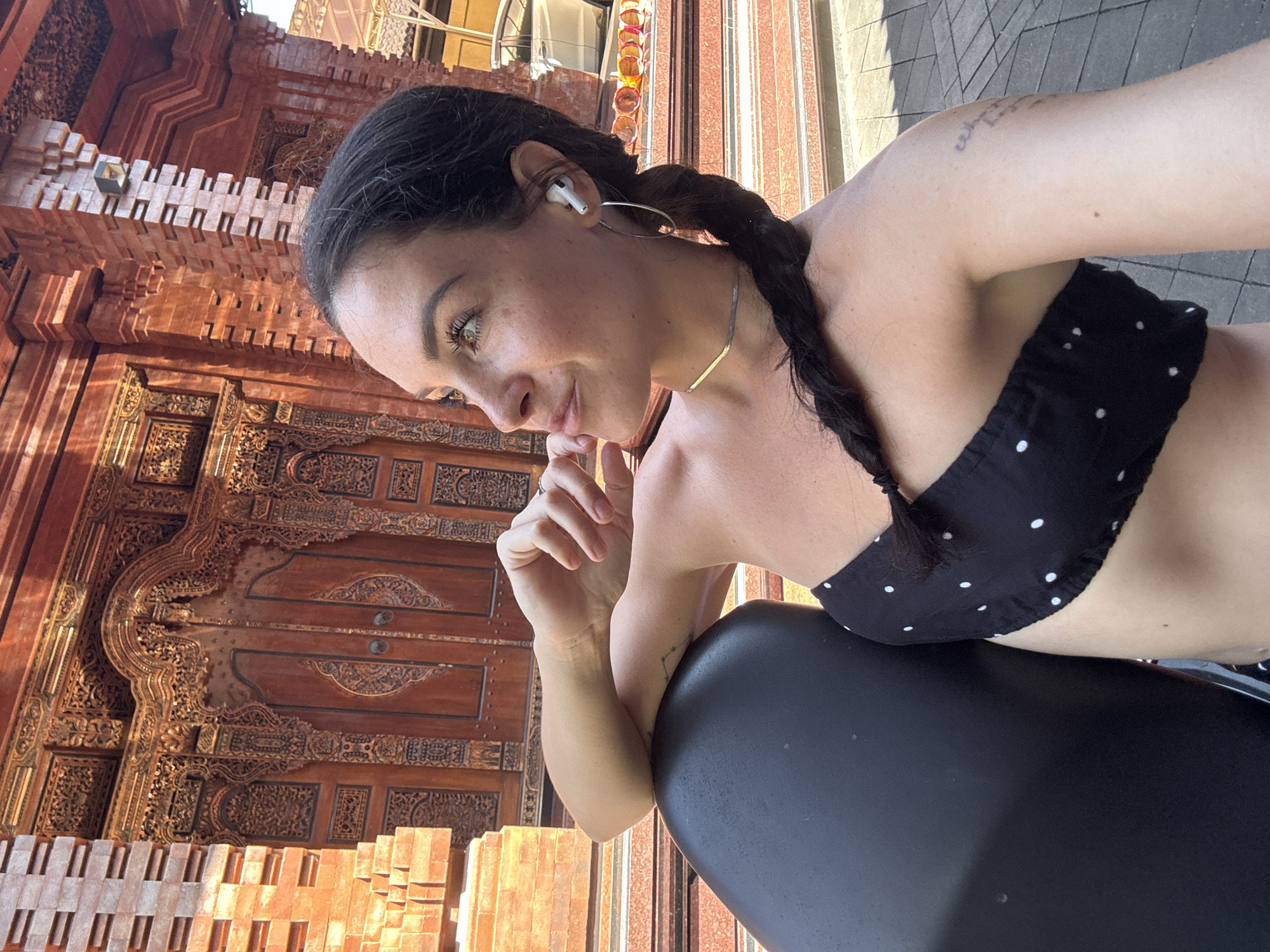 A woman with dark hair in a braid is seen from the chest up, resting her left arm on a dark, curved object. She is looking to her right, with her right hand near her chin. She is wearing a black, polka-dotted strapless top and a thin silver necklace. In her right ear, she has a white wireless earbud, and a large silver hoop earring in her left ear. Behind her, a large, intricately carved wooden door is visible, set within a red brick structure. The architecture suggests a temple or traditional Indonesian building. The background also shows some steps, paved ground, and what appears to be a parked car and sunglasses. The lighting suggests it is daytime, with sunlight casting shadows. The scene is likely in North Kuta, Indonesia, given the architectural style.