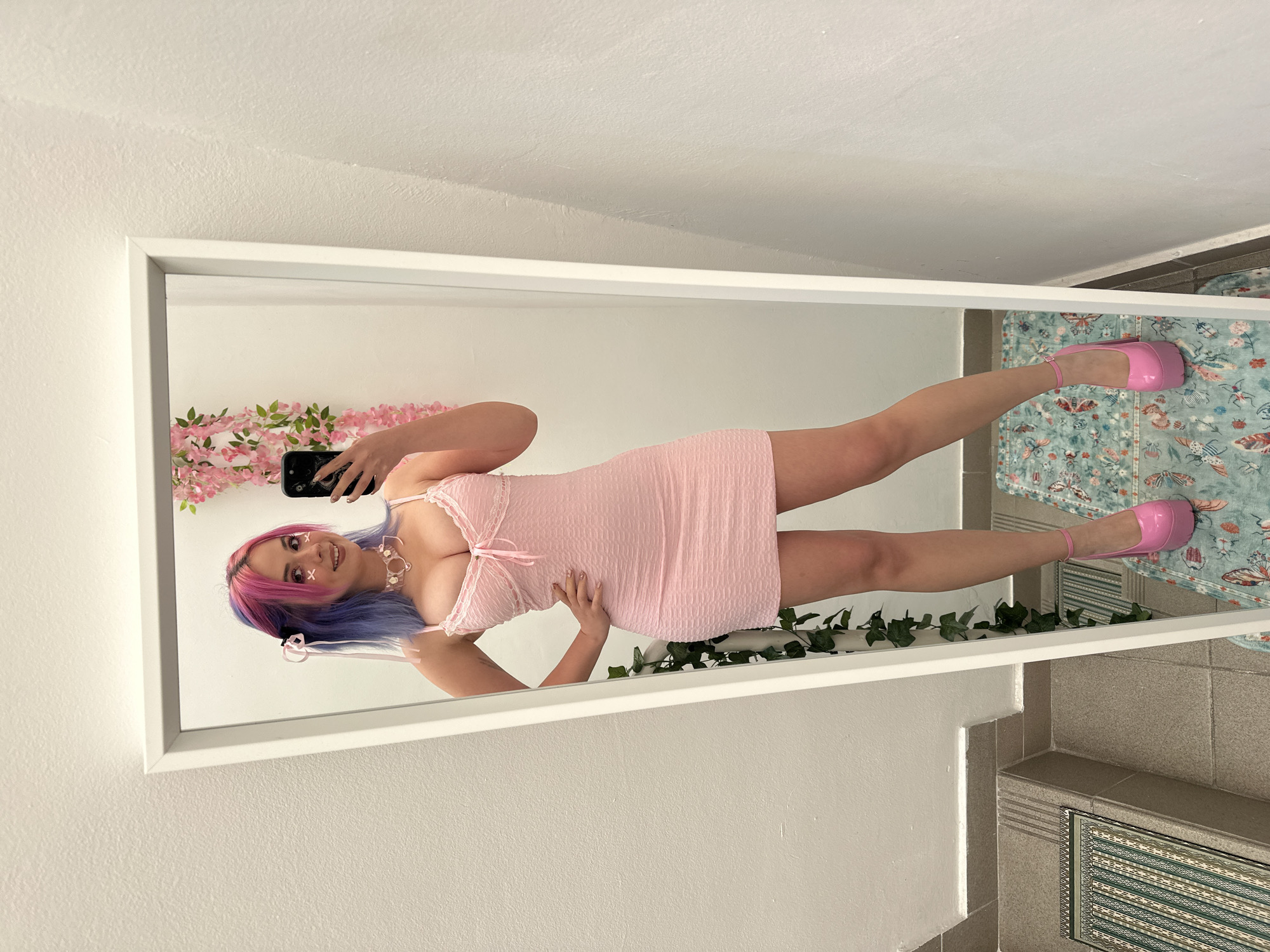 A person with pink and purple hair is taking a selfie in front of a mirror. The mirror is decorated with flowers on top and ivy on the bottom. The person is wearing a pink dress and matching pink platform shoes. The floor and wall are tiled. A rug is placed on the floor. This scene is inside a building in City of Zagreb, Croatia.