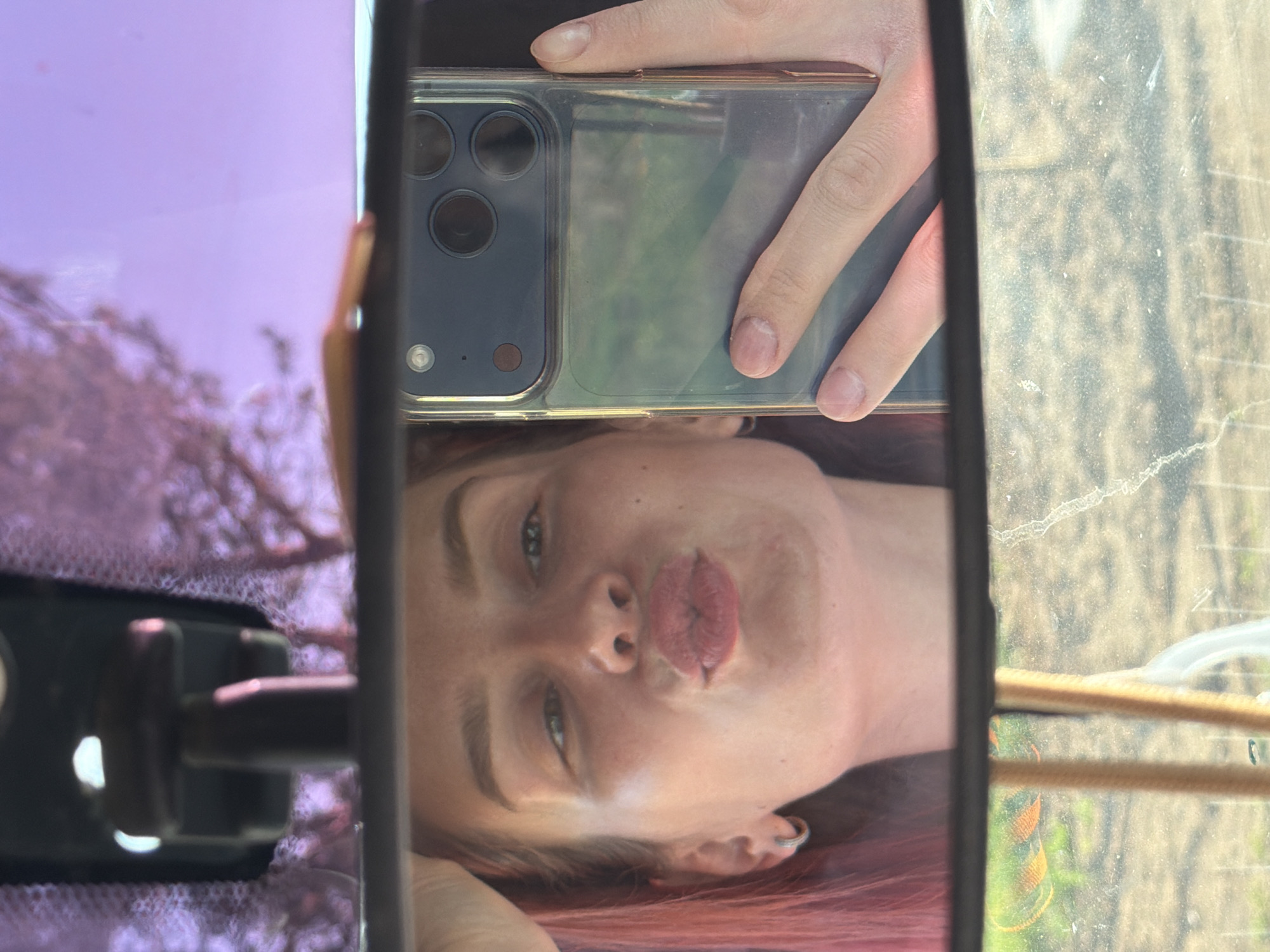 A woman with red hair is taking a selfie in a rearview mirror. She holds a phone with multiple camera lenses. The mirror reflects a tree and a surface possibly covered with snow or ice. The exact location is unclear from the image alone, and it is not possible to determine that this image was taken in Chaikovsky, Russia.