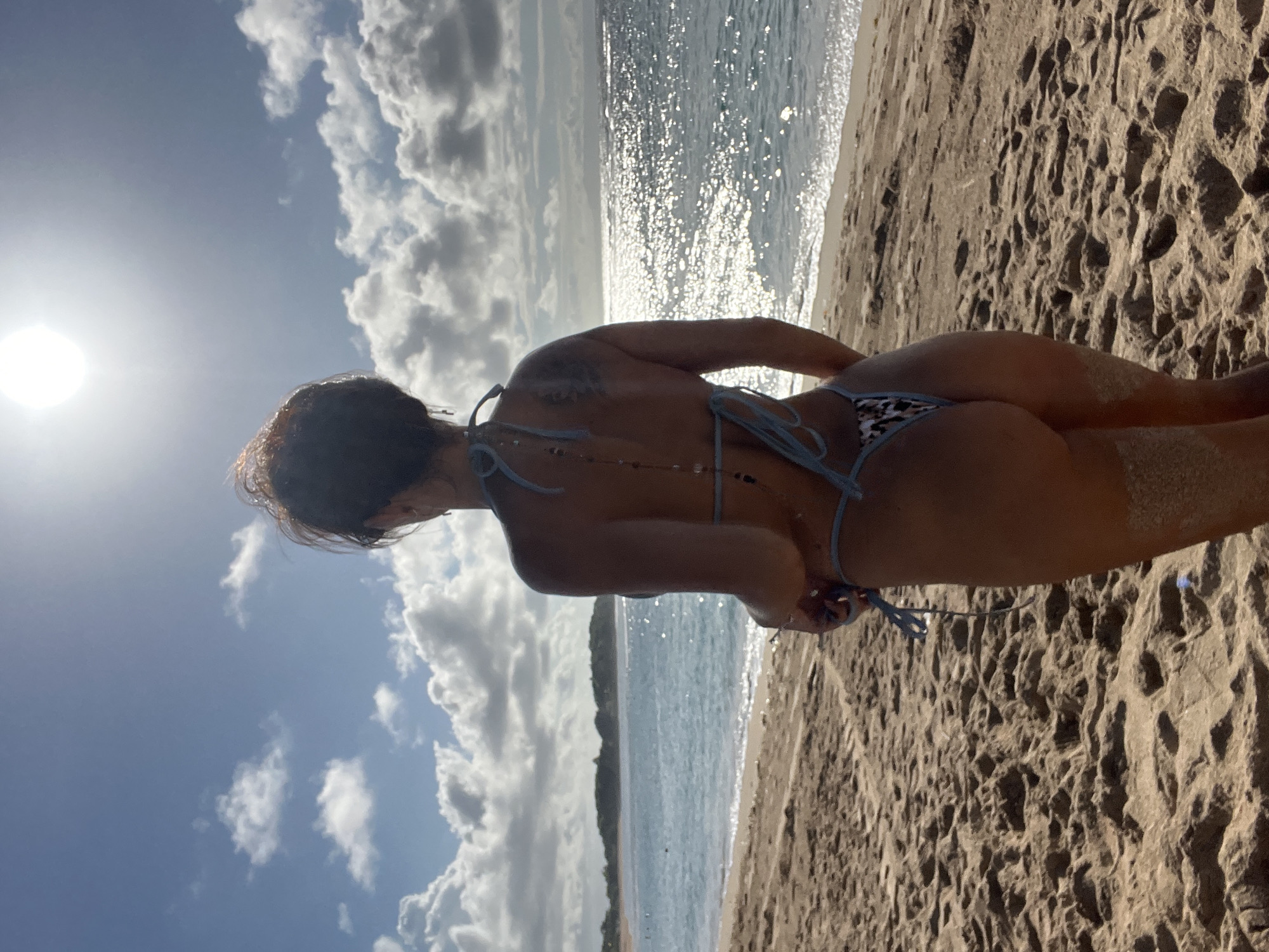 The image captures a woman in a bikini on a sandy beach. The sky is bright, with clouds visible, and the sun is shining. The ocean stretches to the horizon. The photo was taken at Kuta Selatan, Indonesia.