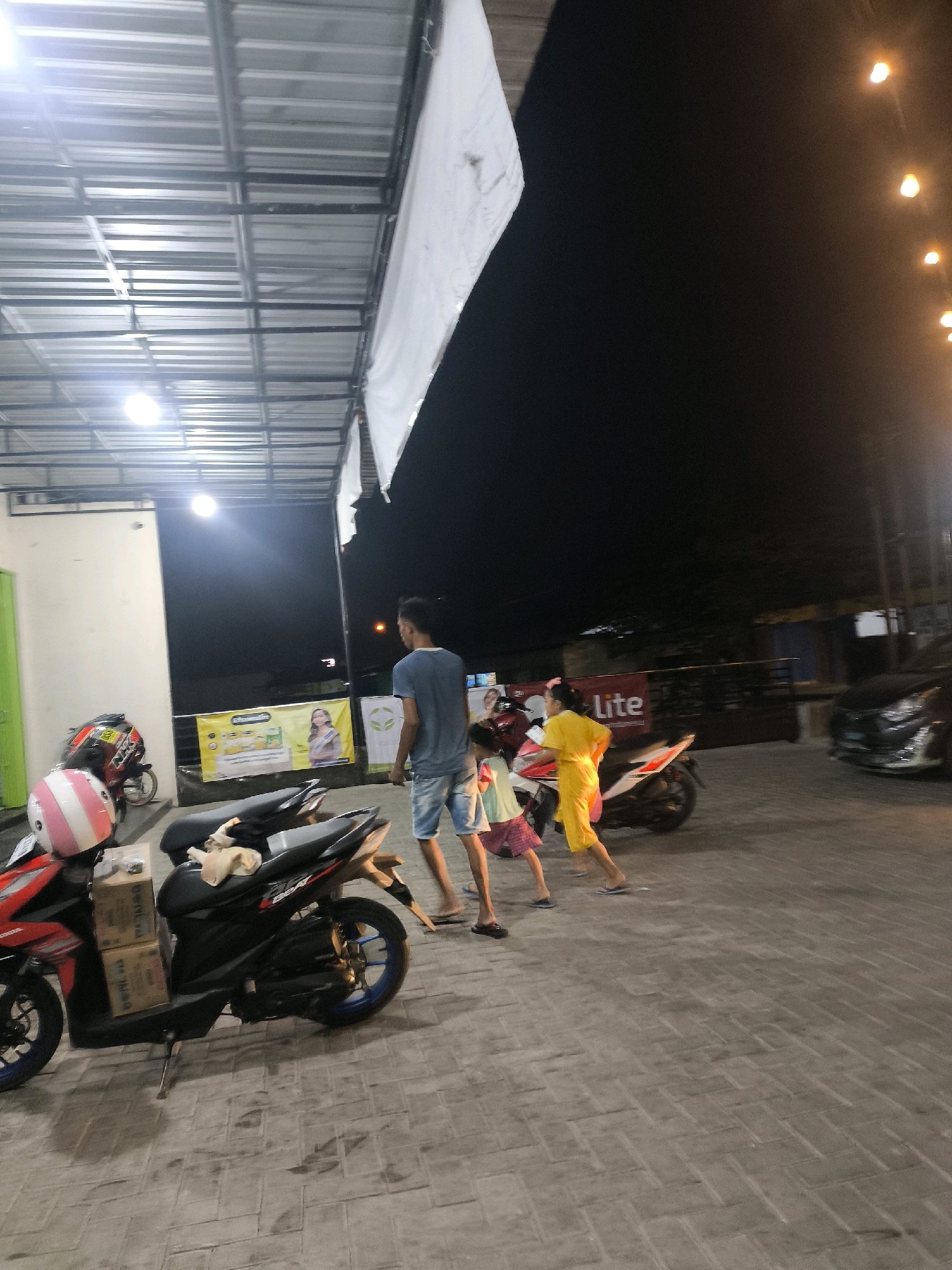 At night in Pilolahuwa, Indonesia, a group of people are gathered near motorcycles. A man in a blue t-shirt and shorts stands facing away from the camera, while two young children, one in a yellow dress and the other in a pink and white striped skirt and a light blue top, are in front of him. A red and black motorcycle with a pink and white helmet and two boxes is parked in the foreground. Another motorcycle is parked behind it. The ground is paved with interlocking bricks. In the background, a banner with a picture of a woman and text is visible, as well as a black car. The scene is illuminated by overhead lights and string lights.