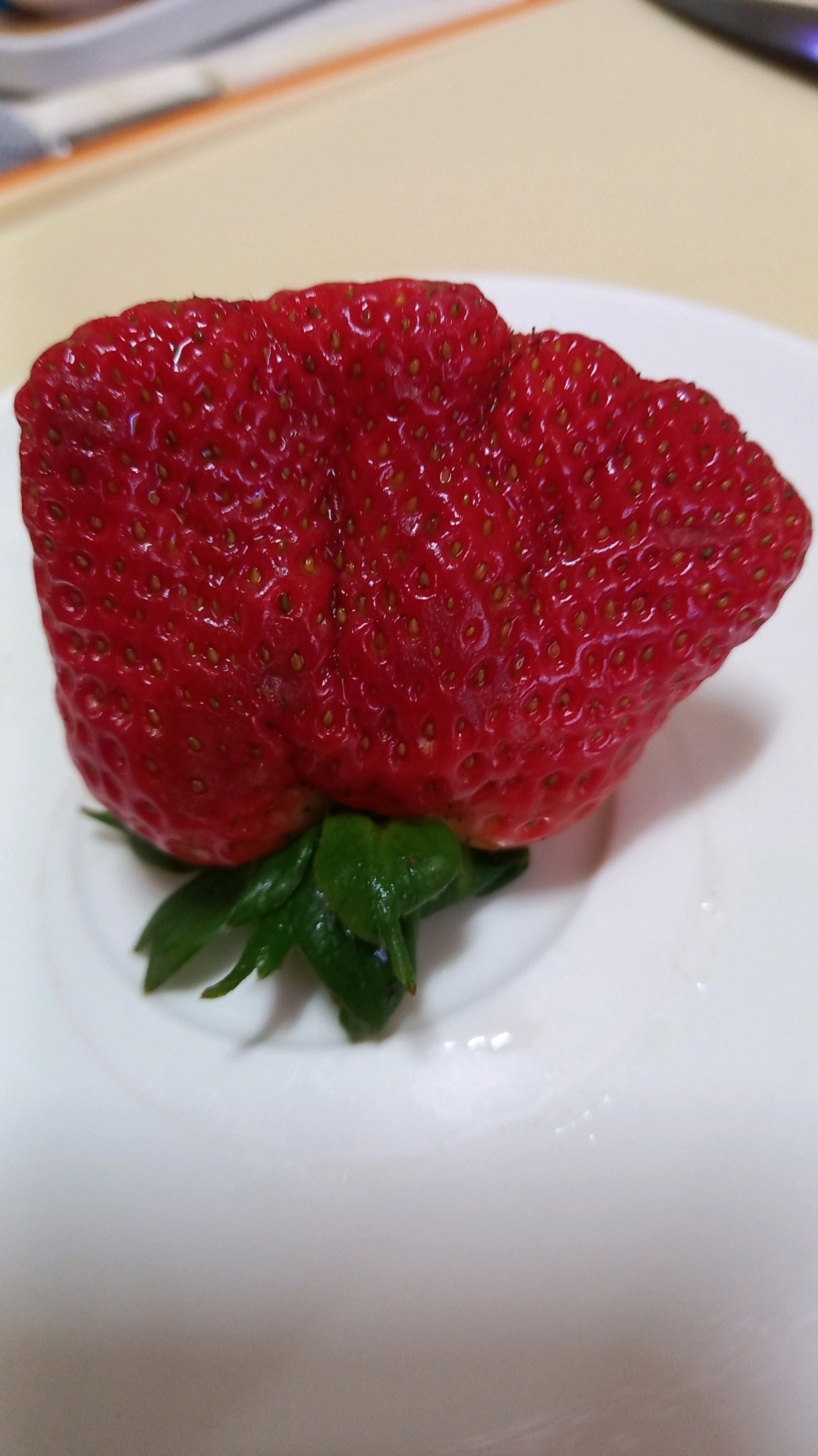A single, large, red strawberry with a textured surface and visible seeds sits on a white plate. Green leaves are attached to the bottom of the strawberry. The background is blurred, showing a light-colored surface with some indistinct objects, possibly indicating an indoor setting. There are no people or animals present in the image. The strawberry's shape is irregular, suggesting it may be a naturally occurring anomaly or a result of specific growing conditions. The lighting is bright, highlighting the sheen on the strawberry's skin. The location in Ko Pha-ngan District, Thailand, is not discernible from the visual information provided in the image.