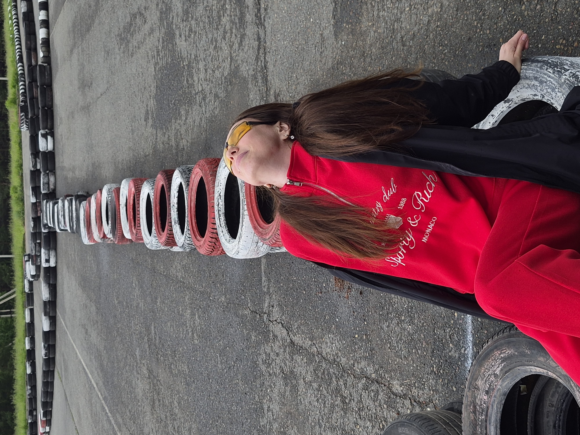 The scene depicts an outdoor asphalt track in Прикамский, Russia, exhibiting surface cracks and wear. A prominent barrier constructed from vertically stacked automobile tires, alternately painted red and white, extends diagonally from the lower right foreground towards the upper center. In the upper background, additional tire barriers, primarily black and white and stacked horizontally, delineate a track turn and a perpendicular section. A lone female individual is positioned in the lower right, leaning backward against the red and white tire barrier. She is dressed in a red tracksuit, featuring white embroidered text on the chest that includes "Sporty & Rich," "Country club," "1988," and "MONACO." A black lightweight jacket is draped open over her shoulders and arms. She wears yellow-tinted sunglasses, and her head is tilted upwards with her neck extended. Her long brown hair falls across her shoulders. Her left hand rests on a white-painted tire section. Several unpainted, dark gray tires are visible in the lower left foreground, alongside the track.