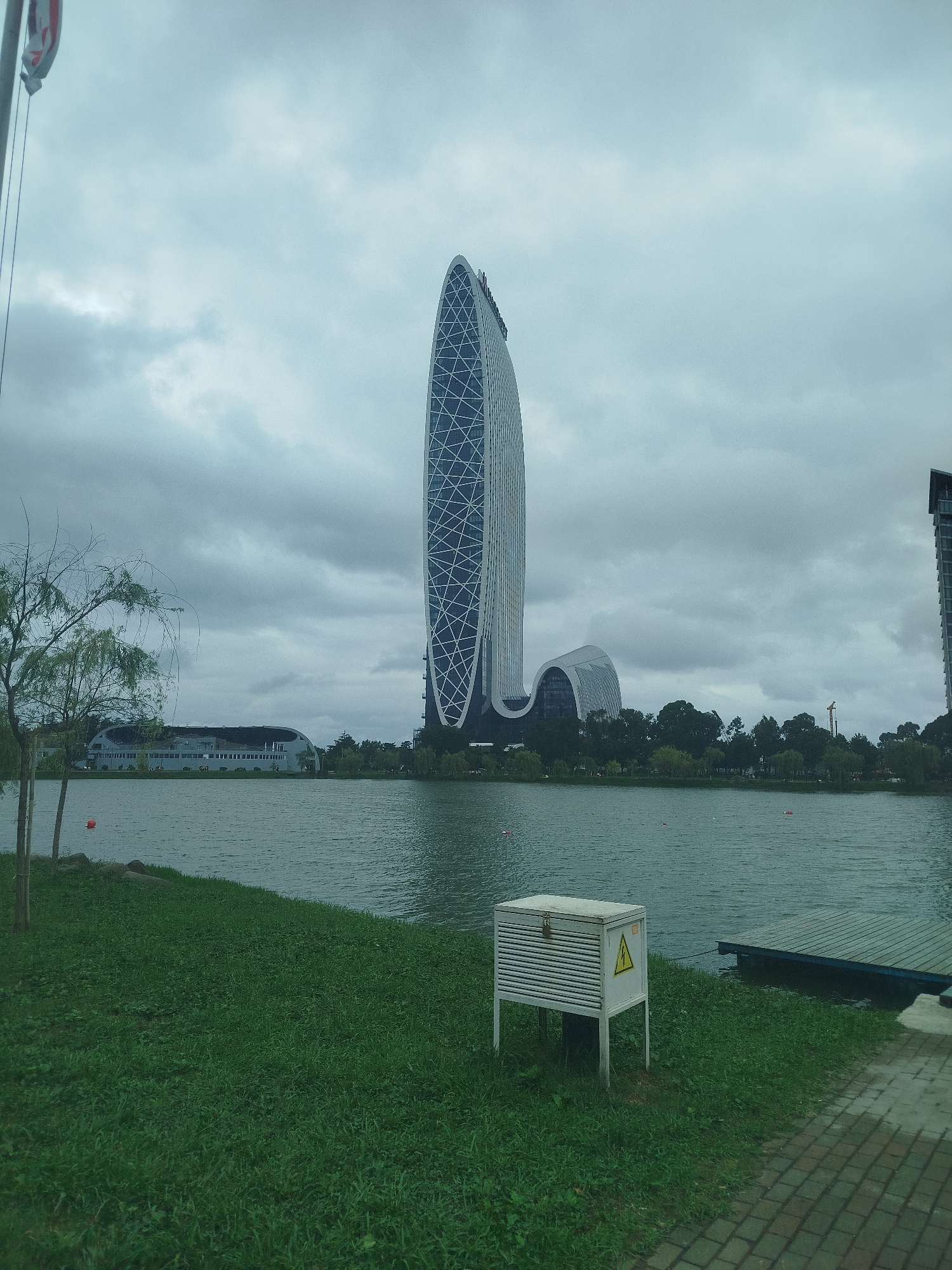 A panoramic shot shows a cloudy day over the New Boulevard area of Batumi, Georgia, featuring the distinctive architecture of the Alphabet Tower and the surrounding cityscape. The scene is dominated by the lake in the foreground, with a grassy embankment leading to the water. A small, orange buoy is visible in the lake near the left bank. On the grass, a grey electrical box with a yellow hazard symbol is positioned. Beyond the lake, a row of trees separates the water from a long, low-rise building with a curved roof, possibly part of the Batumi Technological University. To the right of this building, the Alphabet Tower, a striking modern structure resembling an DNA helix, stands tall. Further to the right, another modern high-rise building can be seen. A wooden dock extends into the lake on the right side of the image. The overall atmosphere is serene, with muted colors due to the overcast sky.