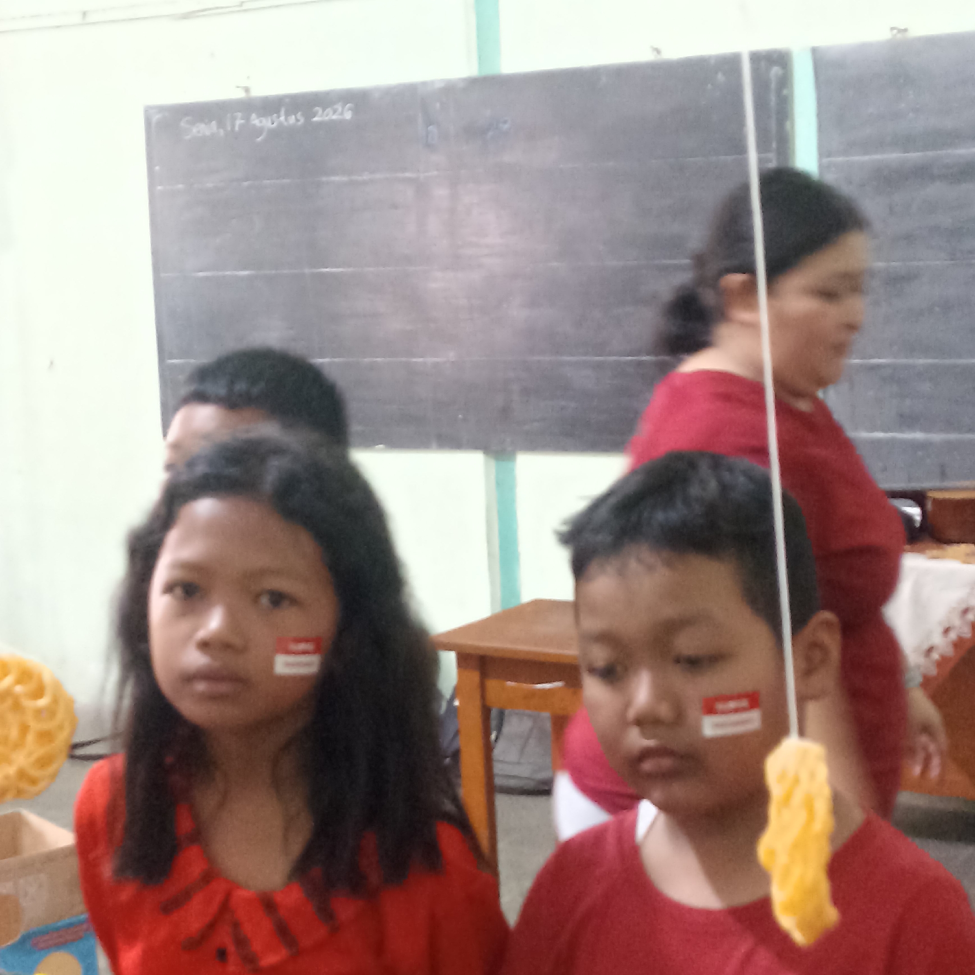 The image is an indoor scene, likely a classroom or community hall, with a blackboard as a prominent backdrop. The blackboard has white chalk writing that appears to read "Senin, 17 Agustus 2026." Three children are visible in the foreground, two of whom are in focus. They are all wearing red shirts. The child on the left, a girl, has black hair and a red and white sticker on her cheek. The child in the center, a boy, also has black hair and a similar red and white sticker on his cheek. He is holding a string with a yellow, puffed snack attached to it. A third child, a boy, is partially visible behind the girl. In the background, a woman in a red shirt is out of focus. A wooden table is positioned to the right of the children. The overall impression is of a festive gathering, possibly related to Indonesian Independence Day given the date and the Indonesian flag colors represented on the stickers.