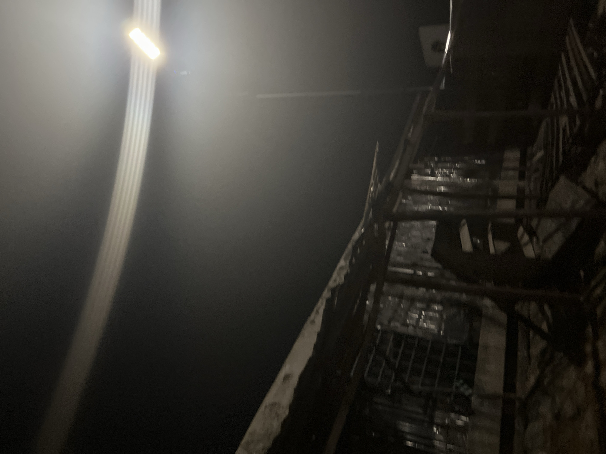 In an outdoor, nighttime scene, a single, bright street light illuminates the area from above and to the right. The light casts a glare across the top of the image, with horizontal lines of light streaking through the dark sky. Below the light, a dark, dilapidated structure is visible, partially obscured by shadows. The structure appears to be made of corrugated metal and wood, with some sections appearing broken or torn. A window with bars is visible on the left side of the structure. The ground in front of the structure is rough and uneven, covered in debris. In the background, a faint building with a square window is discernible, possibly a house or shop, also lit by the street light. The overall impression is one of neglect and decay in an urban environment.