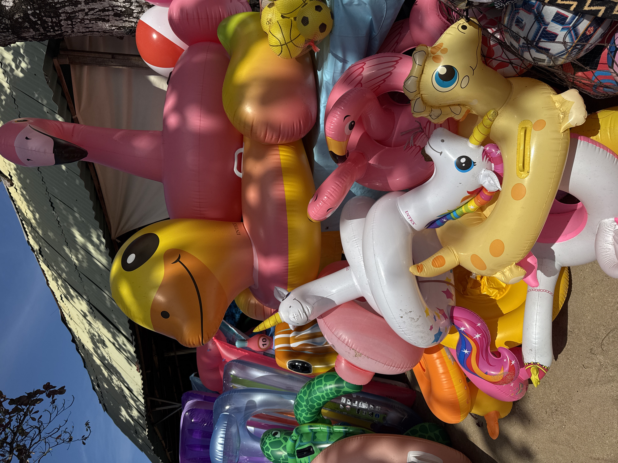 A large, haphazard pile of inflatable pool toys is depicted in an outdoor setting, likely in Kuta Selatan, Indonesia. The pile primarily consists of pool floats shaped like flamingos (both pink and yellow), a giraffe, a unicorn, a turtle, and an assortment of colorful inner tubes. A red and white beach ball and netted soccer balls are also visible. The location seems to be near a structure with a corrugated metal roof, evident from the upper-left portion of the image. The angle of the sun indicates it is daytime, possibly mid-morning or mid-afternoon. There are no people in the image. The setting and the toys suggest a recreational or tourist-oriented location.