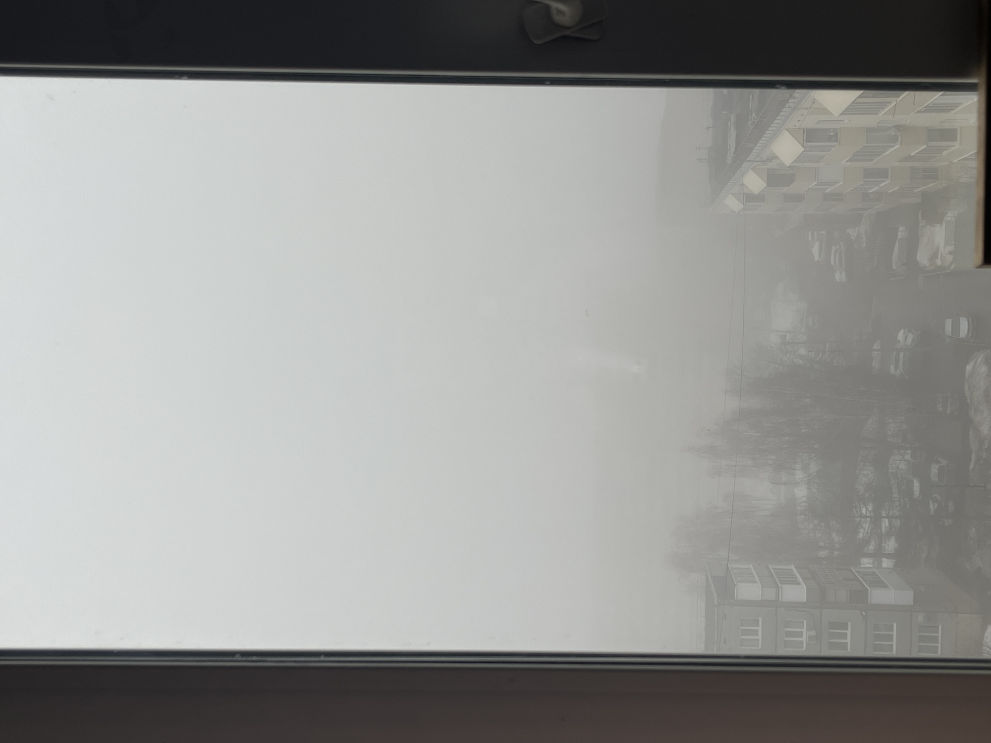 The image shows a view from a window, likely in an apartment building, looking out onto a foggy or hazy cityscape. In the distance are blurred outlines of buildings and trees, which are difficult to discern due to the heavy fog. The window frame is visible in the foreground, indicating an indoor setting. The general atmosphere is overcast and visibility is significantly reduced. No people, specific actions, or interactions are visible. Based on the location information provided (Neftekamsk, Russia), the scene suggests an urban residential area under foggy weather conditions.