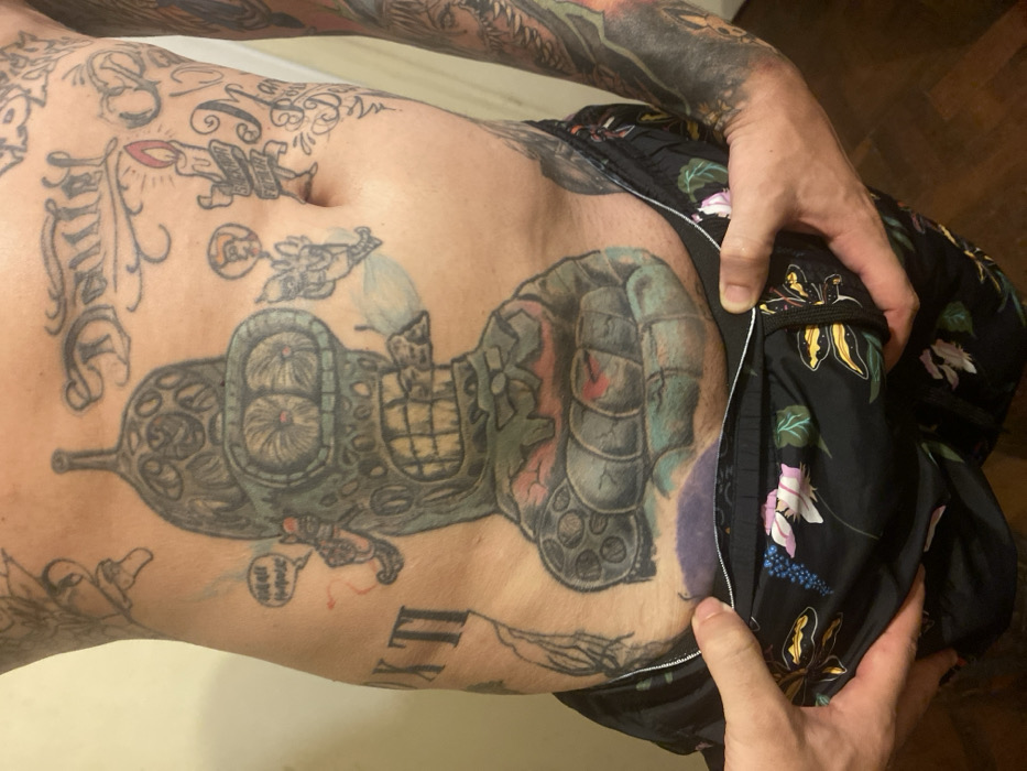 A man with extensive tattoos on his torso and arms is pictured. The largest tattoo on his abdomen depicts a vehicle with steampunk elements. He is wearing floral-patterned shorts, which he is holding open to reveal his side and lower abdomen. His hands are visible, holding the waistband of the shorts apart. The background is a simple, nondescript wall.