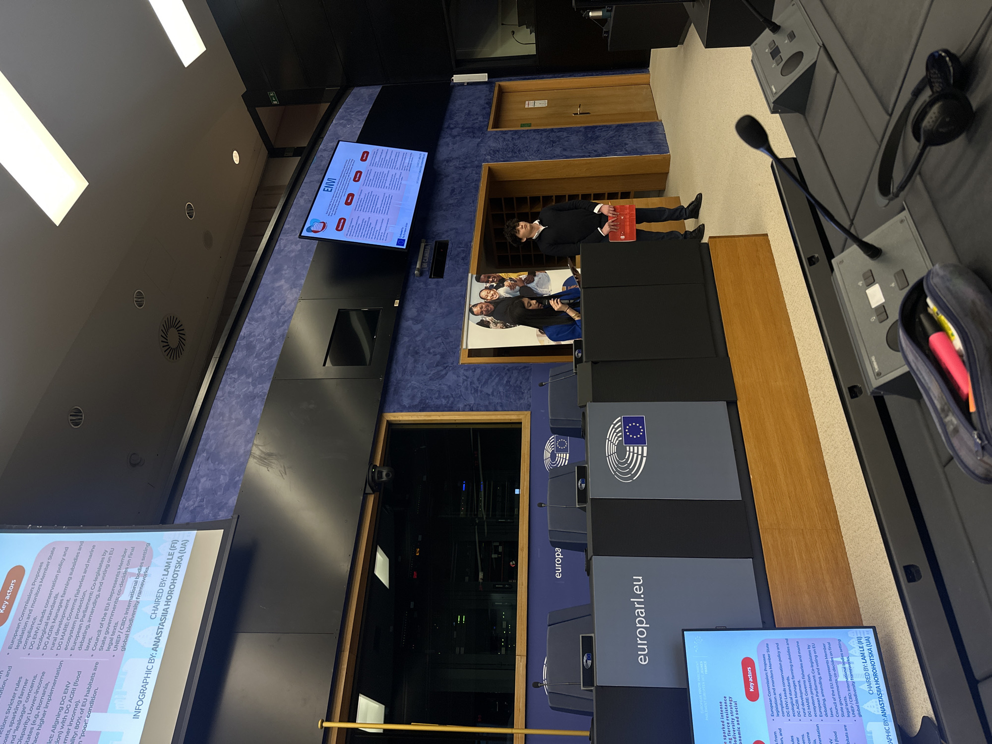 The image shows the interior of a conference room in Strasbourg, France, likely within the European Parliament. The room features rows of desks with microphone consoles and headphones, indicating a formal discussion setting. Large screens at the front display "ENVI" and a slide with information on "Key actors" in the environment sector. A banner "EUROPEAN YOUTH EVENT EUROPÉEN DES JEUNES - FRANCE" is seen. A person in a dark suit stands in a small alcove. There are also other people in the background. A control room is behind a pane of glass with technical equipment. The room is equipped with the European Parliament logo and "europa.eu" branding.