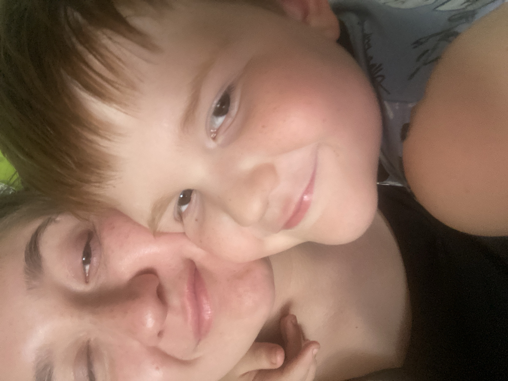 This is a close-up, selfie-style photograph featuring two individuals, likely a mother and her young son, who appear to be in an indoor setting. The woman on the left is looking towards the camera with her left eye closed in a wink. Her skin has a slightly reddish hue, and her lips are slightly parted. She is wearing a black garment. The child on the right is positioned to her right and slightly in front, his head resting on her shoulder. He is looking towards the camera with a slight smile, his eyes visible and his cheeks slightly flushed. His hair is a light brown or reddish-brown color. The background is indistinct and appears to be dimly lit. The image quality is somewhat grainy. There is no discernible indication of the city of Chaikovsky, Russia, or any specific urban environmental features.