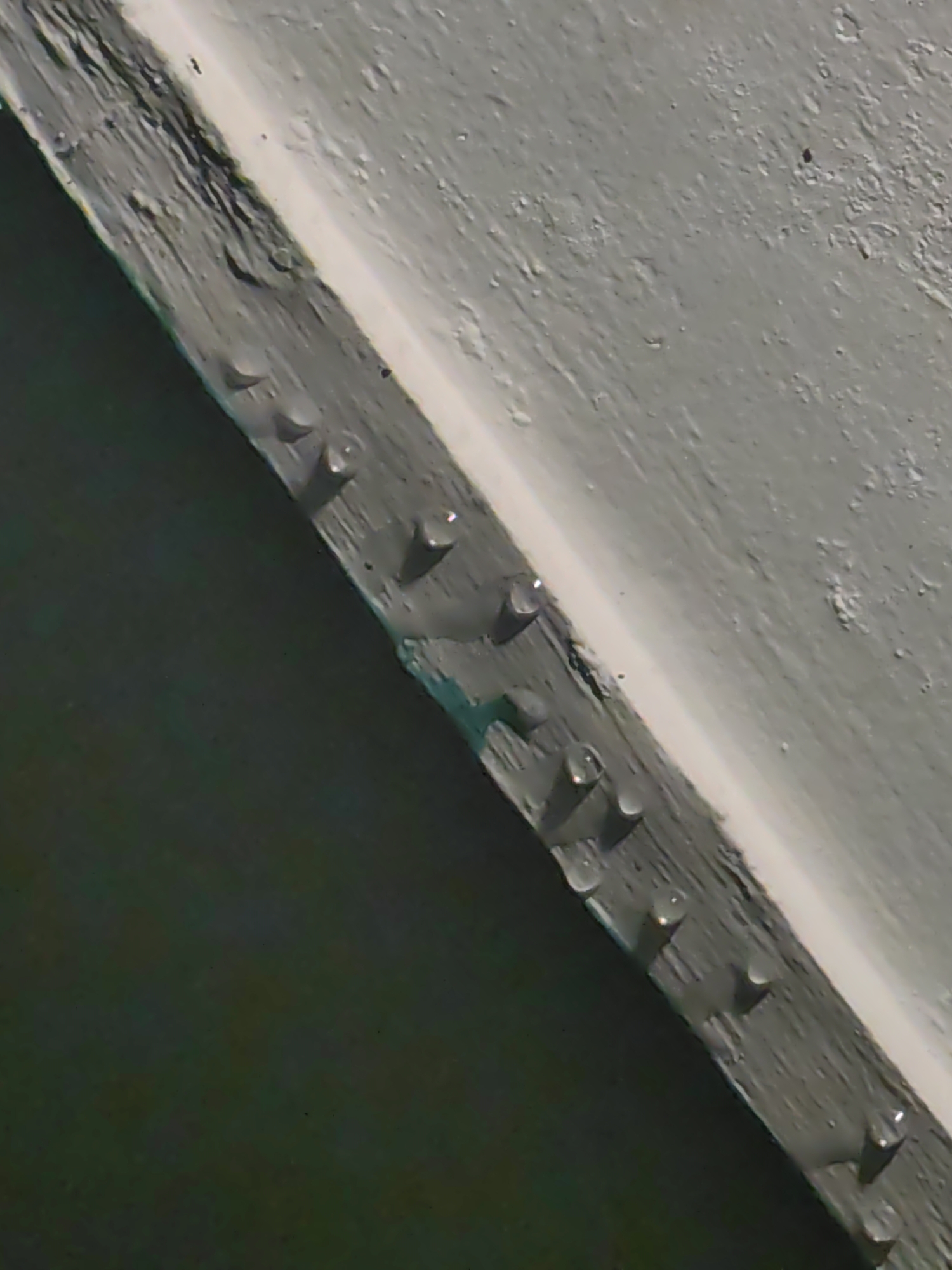 The image is a close-up, oblique view of a textured, grey, weathered surface, possibly wood or concrete, with a lighter colored strip running diagonally from the top left to the bottom right. Several small, round, clear droplets, likely water, are visible on the grey surface, arranged in a line. Some of these droplets appear to be reflecting light. The surface is slightly uneven and shows signs of wear and tear, with some darker green patches visible along the bottom edge of the grey surface. The background is a dark, out-of-focus area. The scene is devoid of any human presence or activity and is likely an outdoor surface in Vashisht, India, given the context.