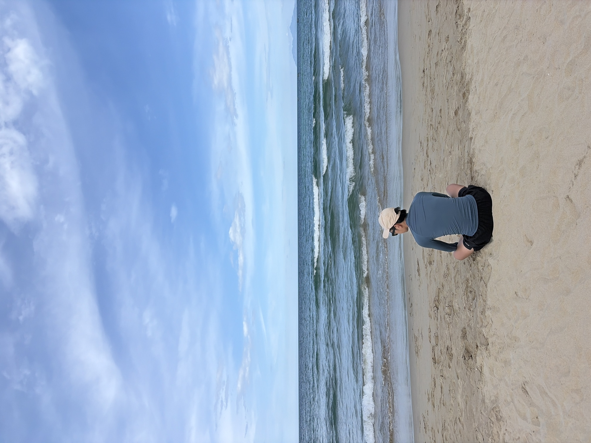 The image depicts a person from behind, sitting cross-legged on a sandy beach in Da Nang, Vietnam. The individual is wearing a long-sleeved blue shirt, black shorts, and a light-colored baseball cap. They are facing the ocean, with gentle waves rolling onto the shore. The sea is a deep blue-green, and the sky above is a clear blue with scattered white clouds. In the distance, the faint outline of mountains can be seen on the horizon. The beach appears to be relatively uncrowded, with the sand showing subtle ripples and textures.