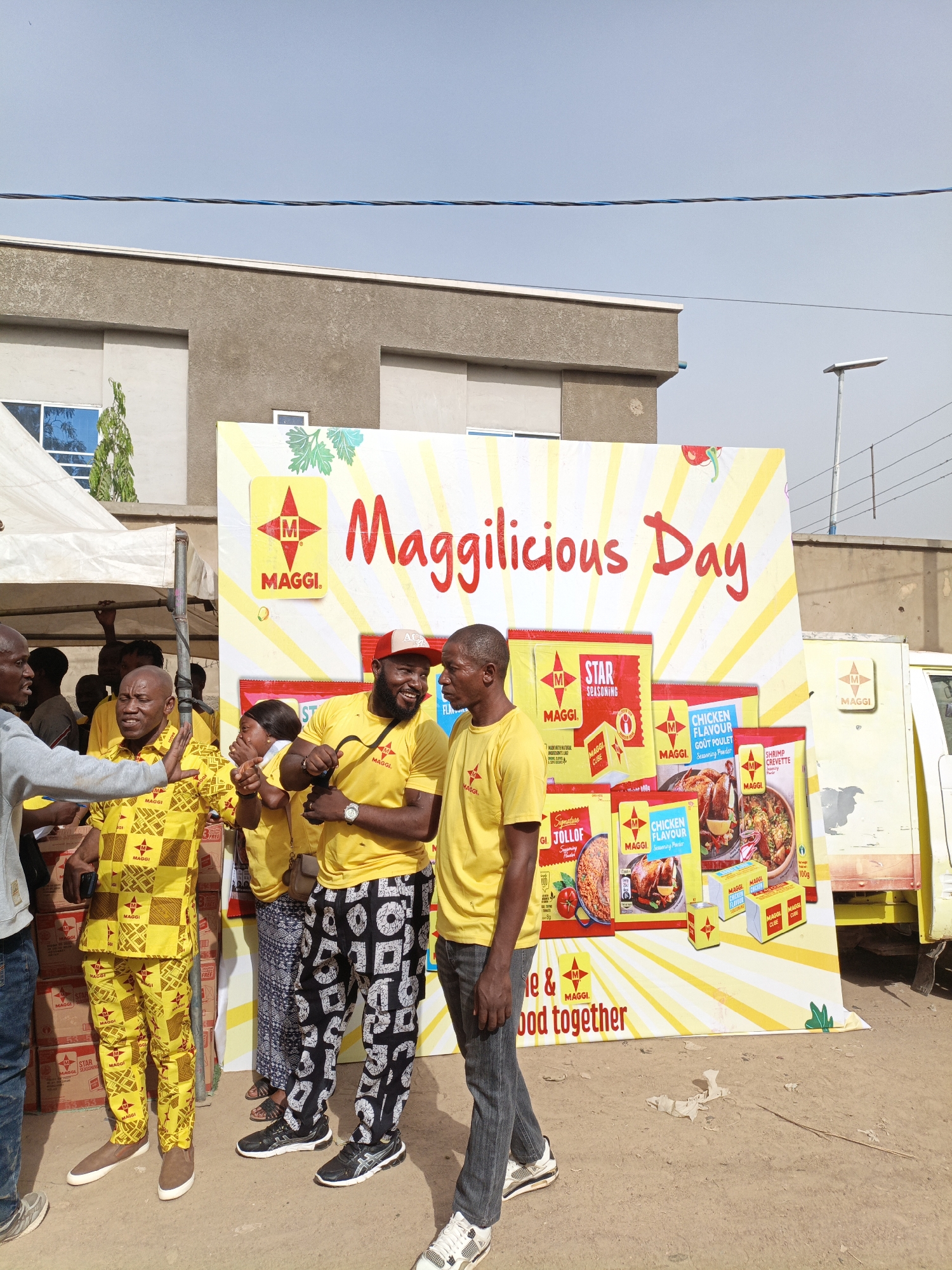 This outdoor daytime scene in Kawaji, Nigeria, depicts a lively "Maggilicious Day" event, likely a promotional gathering for the Maggi brand. A group of adults, mostly men, are gathered, appearing cheerful and engaged in conversation. Many are dressed in yellow attire; some wear Maggi-branded t-shirts, while one man sports a full yellow patterned traditional suit, and another has on a yellow t-shirt paired with black and white patterned pants.
The central focus is a large, brightly colored white and yellow banner that prominently displays "Maggilicious Day" in red script, alongside the distinct "MAGGI" logo. The banner showcases various Maggi products, including "MAGGI STAR SEASONING," "MAGGI CHICKEN FLAVOUR GOUT POULET Seasoning Powder," "MAGGI SHRIMP CREVETTE Seasoning Powder," and "Signature MAGGI JOLLOF," along with Maggi cubes. Text like "MADE WITH NATURAL INGREDIENTS LIKE" and a partially visible slogan "e & bod together" are also present.
To the left of the banner, a white tent or canopy is erected, under which cardboard boxes, likely containing Maggi products, are stacked. On the far right, a yellow and white truck or van, also branded with the Maggi logo, is visible. The ground is a dry, dusty earth. In the background, a building with grey walls and blue-tinted windows stands under a clear, bright blue sky, crisscrossed by overhead power lines. The sunny weather and people's expressions contribute to a festive and community-oriented atmosphere.