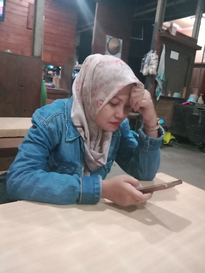 An indoor scene in Kebonsari, Indonesia, shows a woman seated at a light-colored table. She is wearing a light blue denim jacket and a patterned headscarf with shades of beige, pink, and white. Her left hand rests against her forehead, while her right hand holds a brown-cased smartphone, with her thumb positioned over the screen. Red lipstick is visible on her lips, and a metallic-strapped watch is on her left wrist. The background features wooden wall paneling, an artificial light source, a round woven object hanging on the wall, several white plastic bags containing items, a blue bucket, and a yellow object on the floor. The setting appears to be a casual eatery or similar establishment.