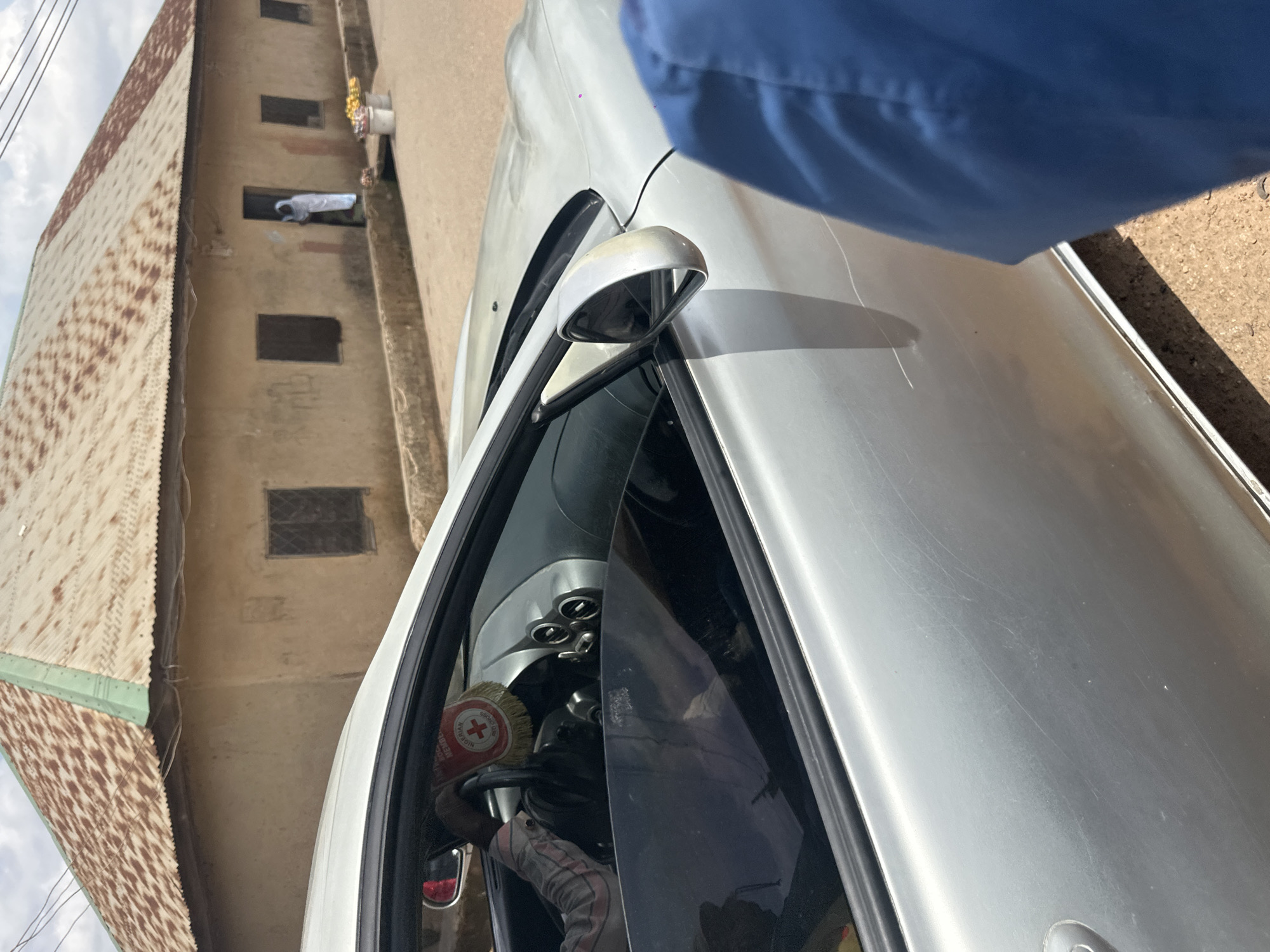 The image shows a person, partially visible, wearing a blue garment, standing next to a silver car. The car's interior, including the steering wheel and dashboard, is visible through the open door. A red and white circular sticker with text is affixed to the steering wheel. In the background, a building with a corrugated metal roof and several windows is visible. One window shows a person in white clothing leaning out. The scene appears to be outdoors, with a cloudy sky overhead. The overall impression is of a casual, everyday scene. The location is identified as Jos, Nigeria, suggesting an urban or semi-urban environment.