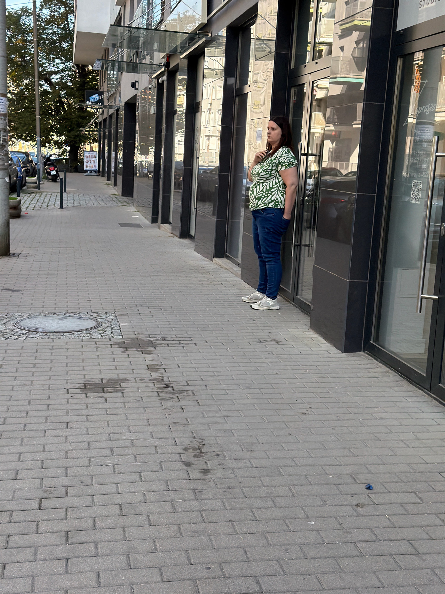A woman stands on a paved sidewalk outside a building in Wrocław, Poland. She is facing left and looking towards the street. She is wearing a green and white patterned shirt and blue jeans. The building appears to be a commercial establishment with large glass windows. The sidewalk is made of gray pavers. In the background, there are trees, a parked motorcycle, and other parked vehicles. The scene is illuminated by daylight.