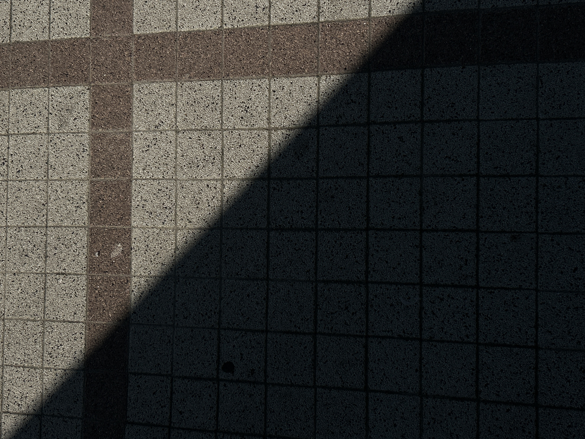 The image depicts a close-up, overhead view of an outdoor tiled surface. The tiles are square and appear to be made of a rough-textured material, possibly concrete or stone, with small flecks of darker material embedded within them. The surface is divided by a grid pattern, with some tiles being lighter gray and others a reddish-brown. A diagonal shadow cuts across the frame, obscuring a portion of the tiles and creating a stark contrast between light and dark areas. The shadow suggests the presence of an unseen object casting it, such as a building or a structure. There are no people, animals, or discernible objects present in the frame other than the tiled ground and the shadow. The environment appears to be a public outdoor space, likely a walkway or plaza, in Tiraspol, Moldova, given the context provided.