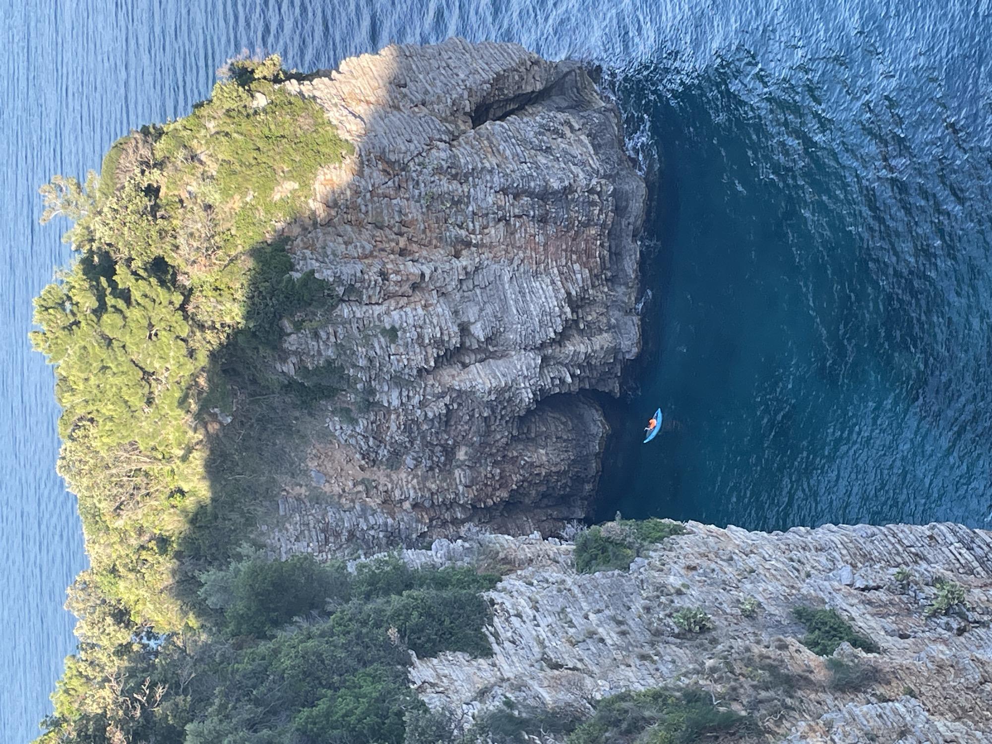 The image presents an aerial view of a coastline in Budva, Montenegro. A rocky cliff formation dominates the central portion of the image. The cliff face descends into deep blue water where a person is visible in a bright blue kayak wearing an orange life vest. The cliff's texture is striated and layered, suggesting sedimentary rock. Vegetation clings to portions of the cliff. The water's surface is slightly rippled. The scene is bathed in sunlight, casting shadows that define the landscape.