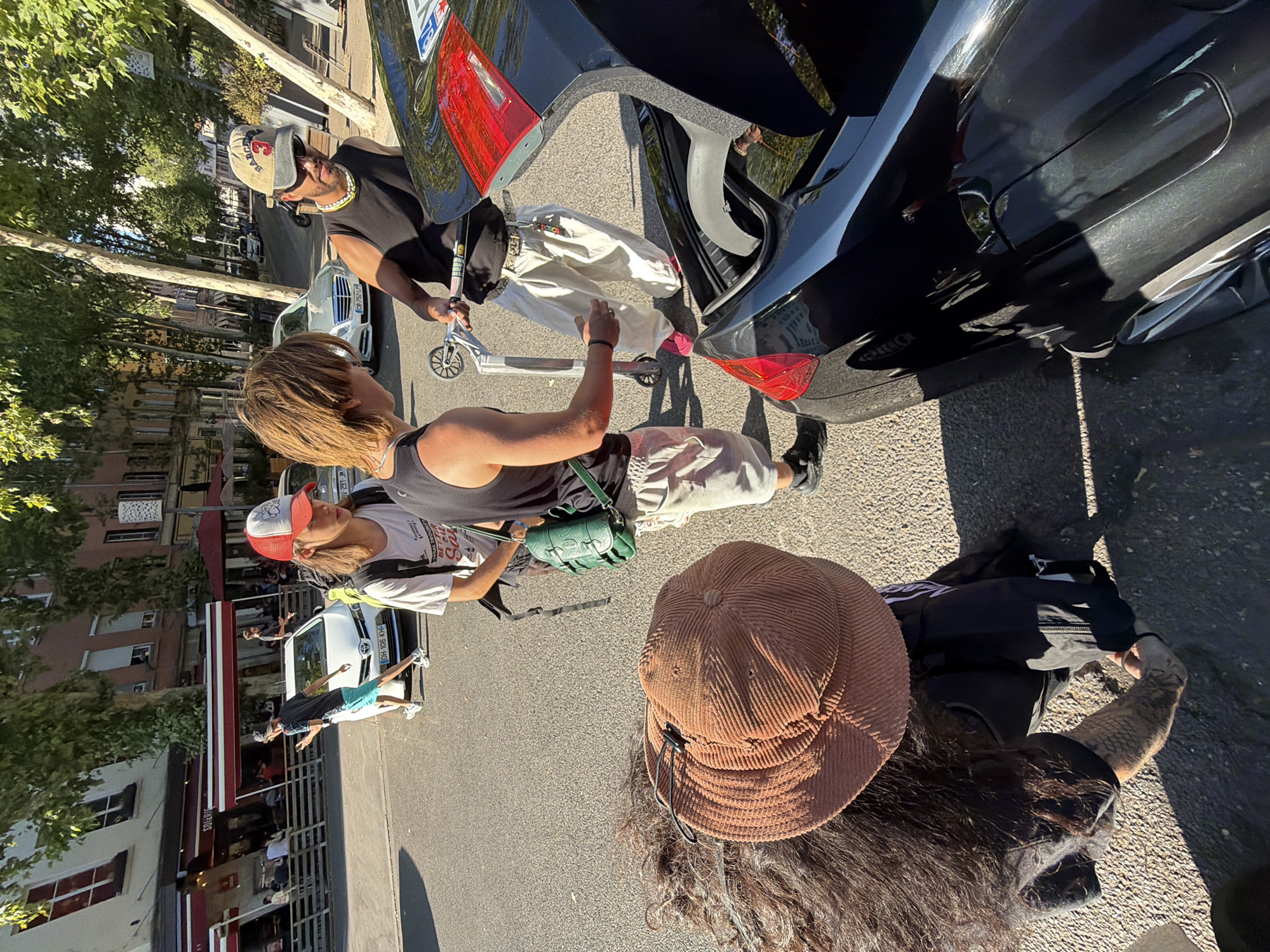 Four individuals are gathered near the open trunk of a black car on a city street. In the foreground, a person with long dark hair and wearing a brown corduroy hat is bent over, with their arm extended towards the car trunk. To their left, another person is partially visible, also bent over. To the right of the car trunk, a person wearing a black tank top and light-colored pants is holding a silver and pink scooter. They are facing towards the back of the car and appear to be interacting with the person holding the scooter. This individual is wearing a tan baseball cap and sunglasses. In the background, another individual in a white t-shirt and a red and white baseball cap is standing. They are holding a green bag. A white car is parked on the street behind them. The scene is set in a city with buildings and trees lining the street, suggesting an urban environment. The lighting indicates it is daytime.
The image appears to have been taken on Rue des Pénitents, near the intersection with Rue de la Mosson, in the 4th arrondissement of Lyon, France. The buildings in the background, particularly the multi-story structures with awnings and visible windows, are consistent with the architecture in this area. The presence of trees lining the street is also characteristic of the neighborhood.