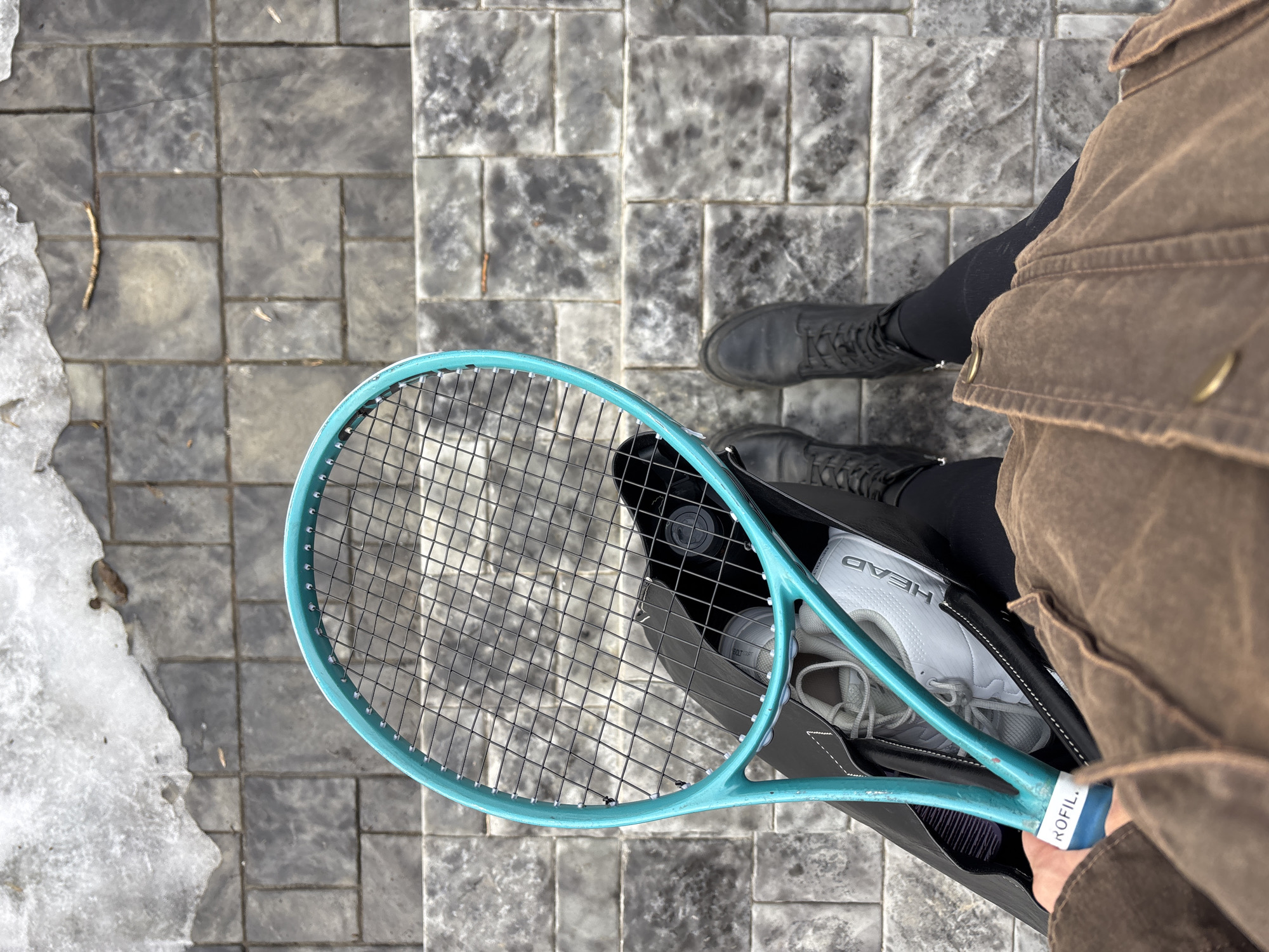 An overhead view captures a person standing on a dark grey paver sidewalk in Chaikovsky, Russia. The individual wears a brown jacket with visible stitching and brass-colored buttons, black leggings, and black laced boots. The person's left hand holds a teal/aqua blue tennis racket, partially showing "KOFIL" on its grip, along with a black open-top bag. Inside the bag, a pair of white athletic shoes displaying the "HEAD" logo, a black cylindrical object, and a dark comb are visible. A patch of melting snow and ice occupies the upper left portion of the paved area, with small twigs and debris present.
