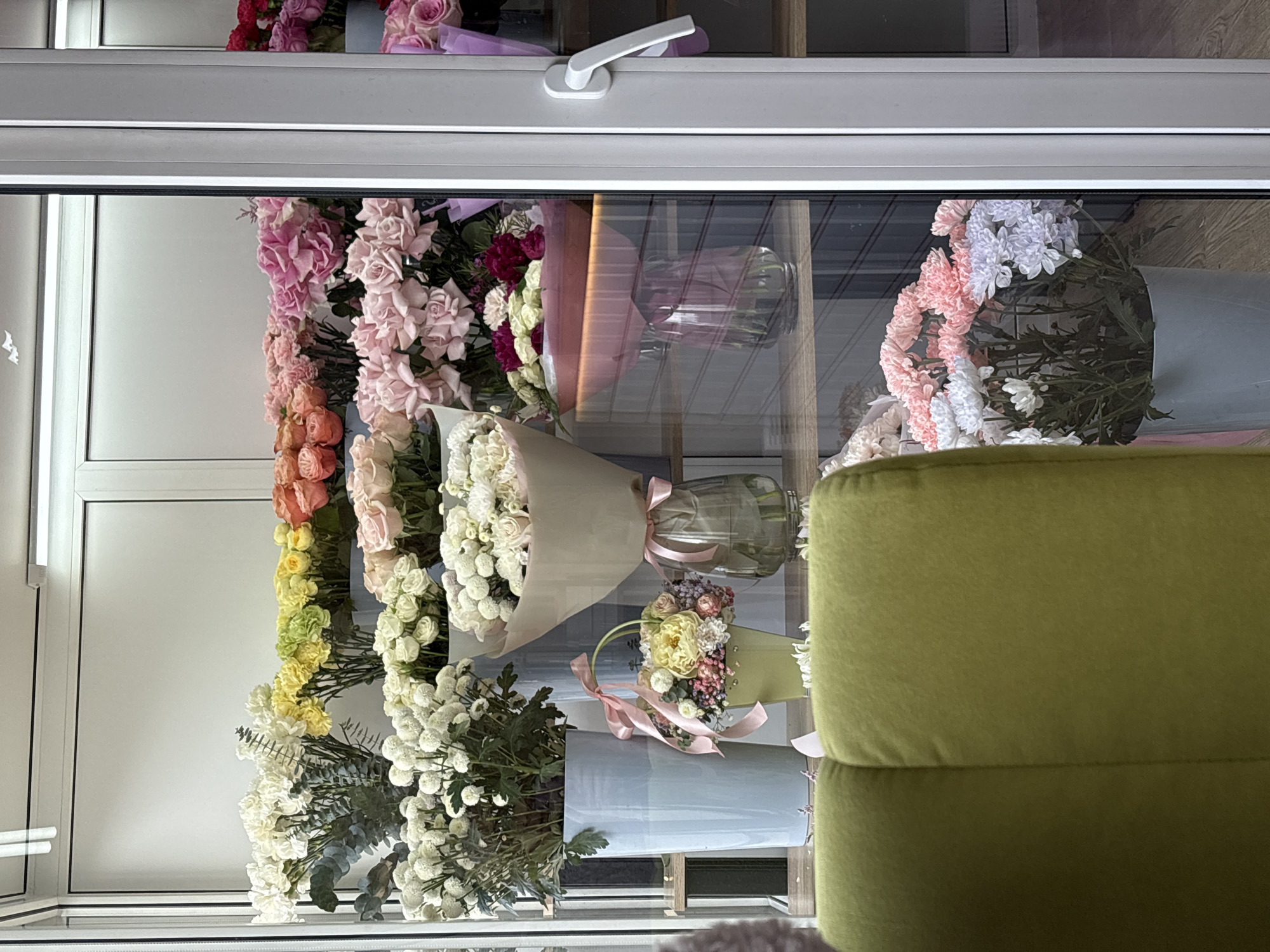 The image shows a display of floral bouquets and arrangements behind a window, possibly a flower shop, in Krasnoyarsk, Russia. The arrangements are of different colors, styles, and contain a variety of flower types including roses, chrysanthemums, and other decorative elements like ribbons and vases. The bouquets are displayed vertically, suggesting they are arranged in shelves. A green piece of furniture, possibly a chair, is partly visible in the reflection on the glass. The window has a white frame and handle. The reflection reveals what might be the interior of a room with wood paneling in the background.