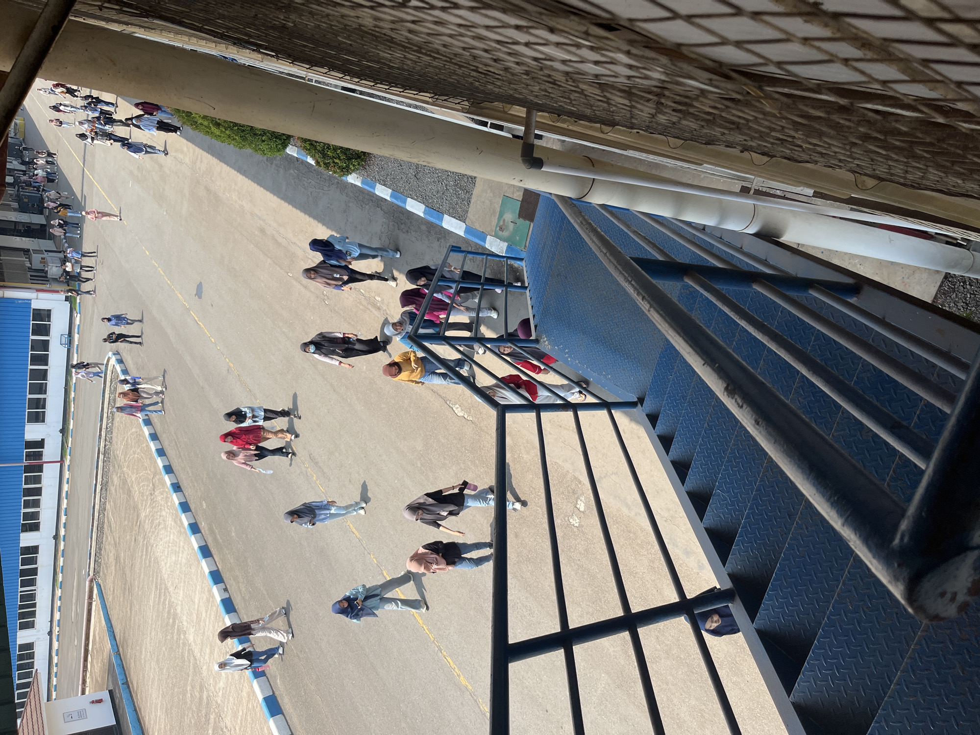 A group of women are walking on a paved area outside a large industrial building. The building has a blue and white facade and is bordered by a blue and white striped curb. The women are mostly wearing hijabs and casual clothing. Some are walking individually, while others are in small groups. One woman is walking down a blue metal staircase in the foreground of the image. The scene appears to be in an industrial complex, potentially in a city like Pelang, Indonesia, given the context provided, characterized by its open paved areas and large buildings. The lighting suggests it is daytime, with shadows cast on the ground.