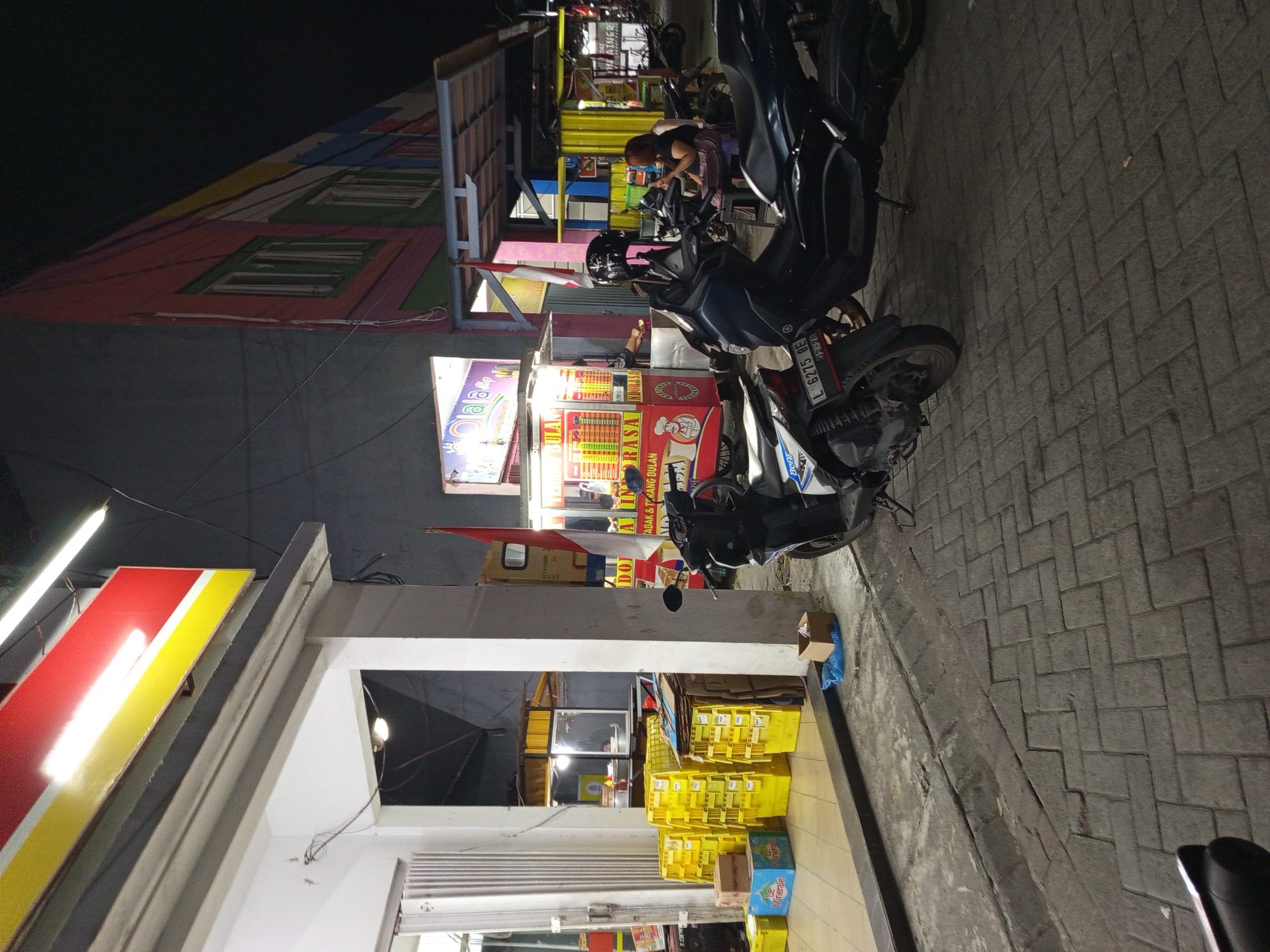 This is a nighttime, eye-level, medium shot of a street scene in Gemurung, Indonesia. In the foreground, two motorcycles are parked on a brick-paved sidewalk. The motorcycle on the left is partially visible, showing its license plate which reads "L 6275 BE". The motorcycle on the right is a dark color and has a helmet on its handlebars. A woman in a dark shirt and patterned skirt is seated on a stool behind the motorcycle on the right, looking at her phone.
To the left of the motorcycles, there is a storefront with a white pillar and a large red and yellow sign with fluorescent lighting. Yellow crates are stacked outside the storefront. In the middle ground, there is a food stall with a colorful sign displaying food items and prices. An Indonesian flag is attached to a pole in front of the food stall. Further back, the facade of a building with colorful vertical stripes and windows is visible. The overall environment suggests a commercial area at night, possibly a street food or small business district.