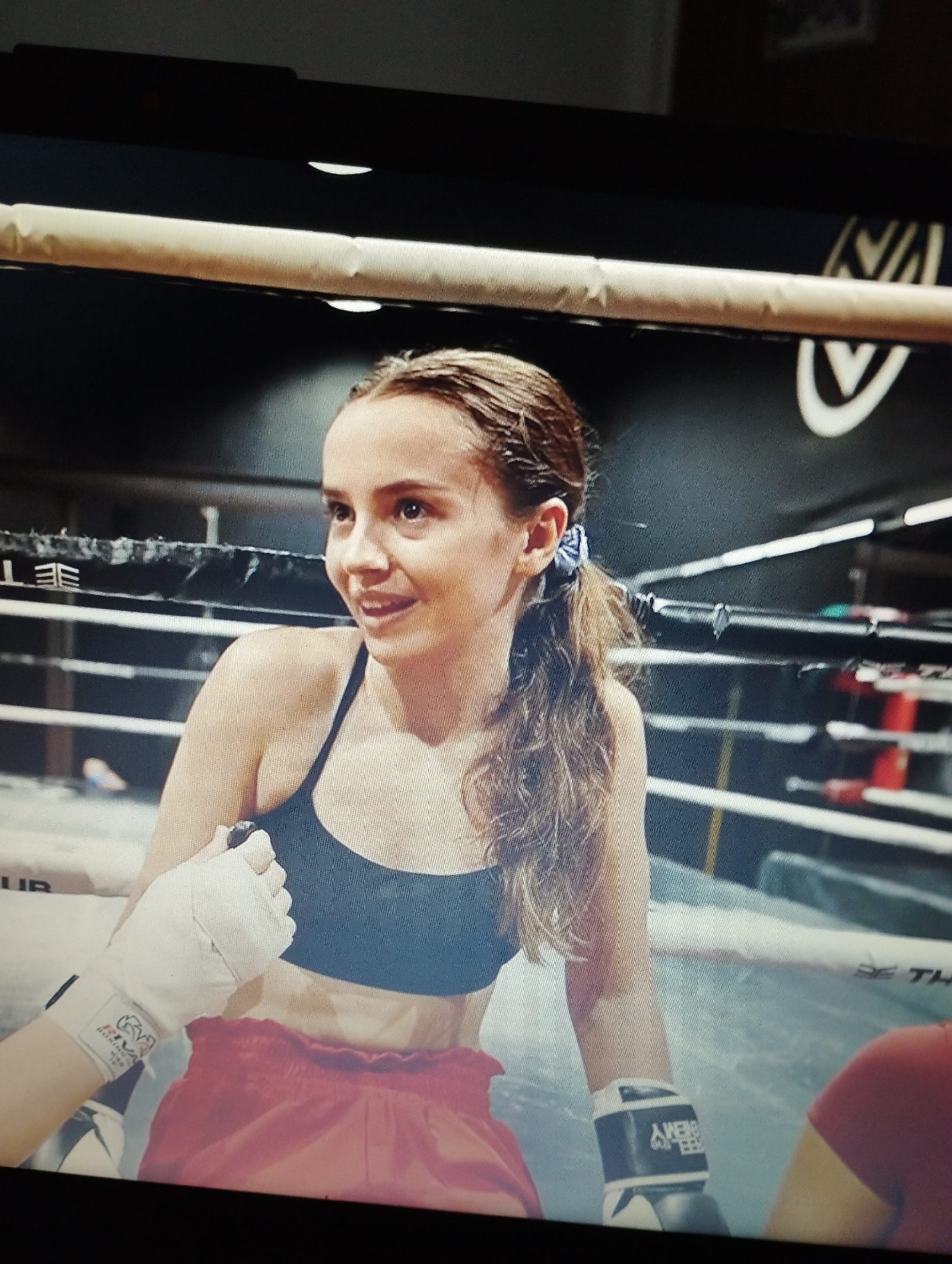 A young woman with braided brown hair pulled back in a ponytail is inside a boxing ring. She is wearing a black sports bra and red boxing shorts. Her left hand is wrapped in white tape, and she is wearing a black and white boxing glove on her right hand. The woman is looking to her left and smiling. The boxing ring has ropes and a mat, and the background is dark with some blurred lights. There is no indication of Santa Ana, Venezuela in the image.